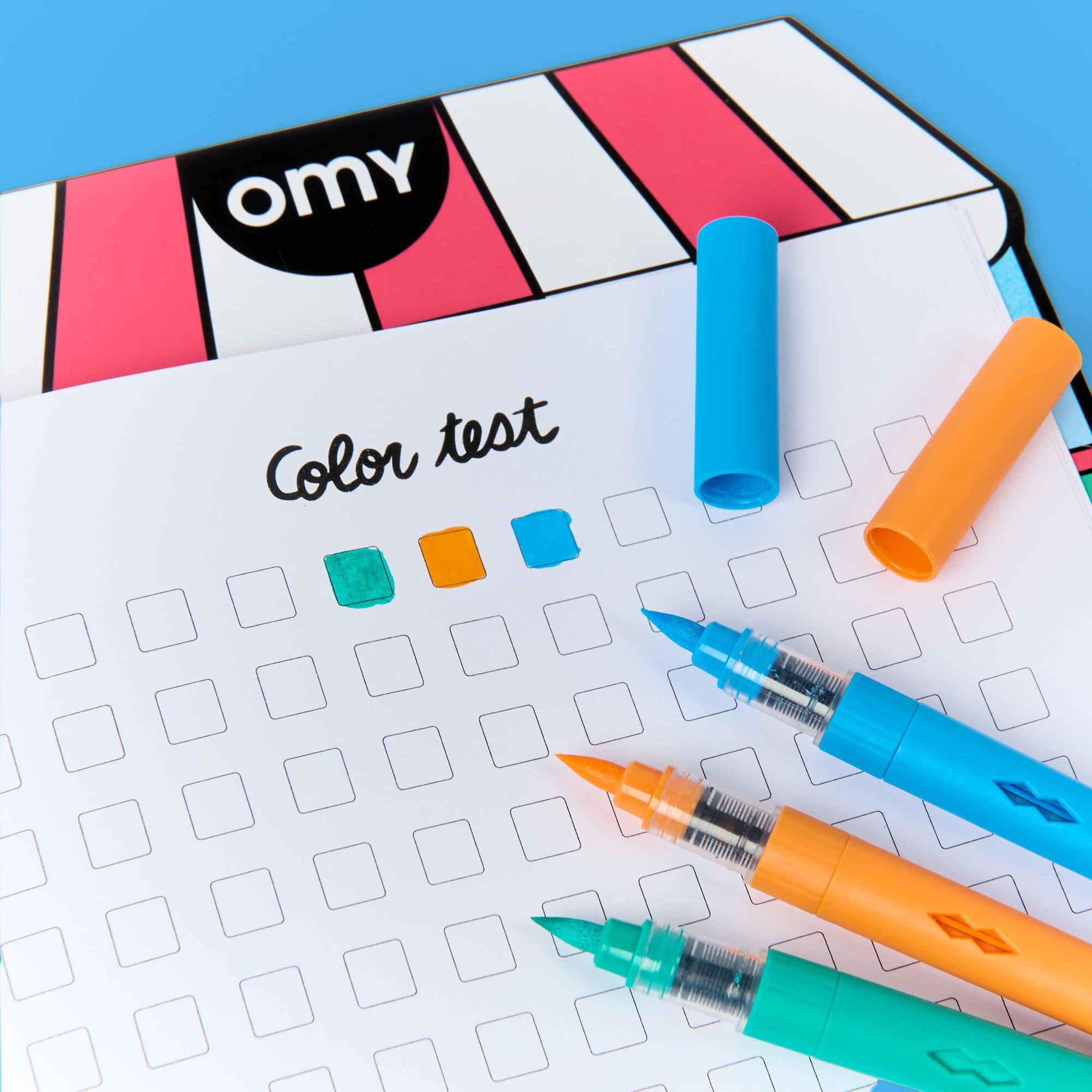 A color test chart with a grid and colored swatches, styled with three markers in blue, orange, and green on a bright blue background. A striped box with the brand name 'omy' is partially visible in the top left.
