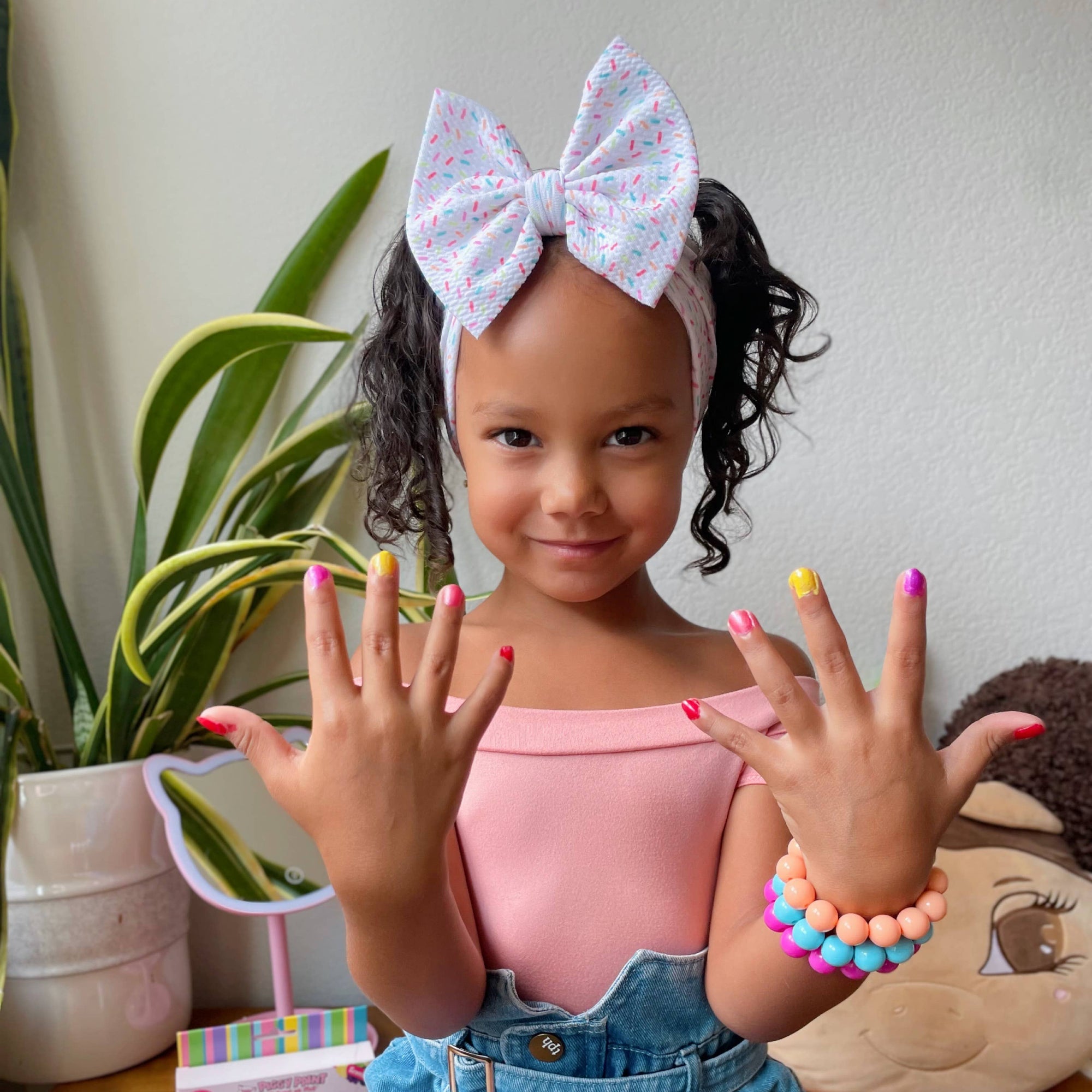 Young girl showing off colorful nail polish with a headband and bracelet, surrounded by nail polish bottles and a plant.