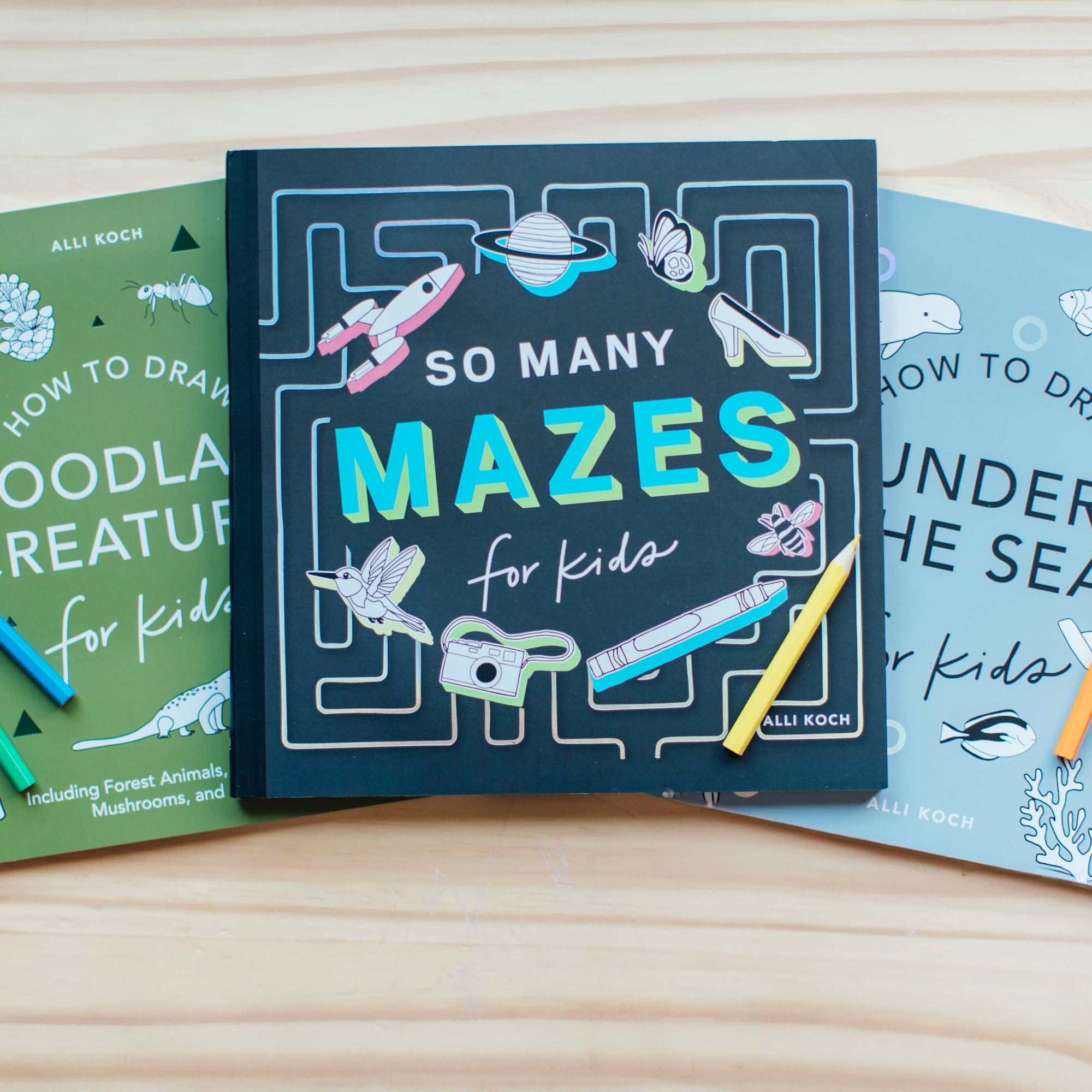 Three children's activity books on a wooden surface with pencils.