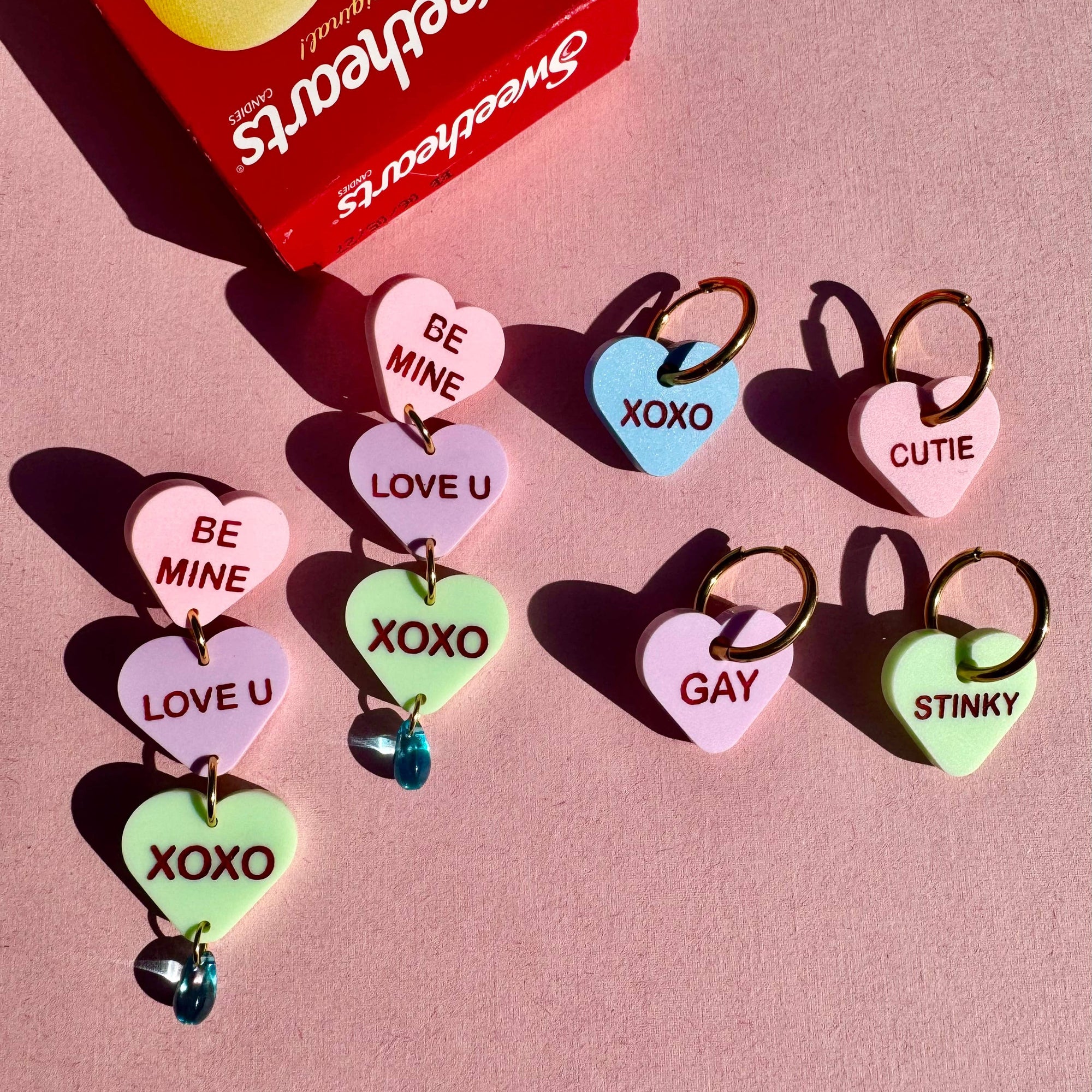 Heart-shaped candy charms with romantic text on a pink background, featuring a Sweethearts candy box.