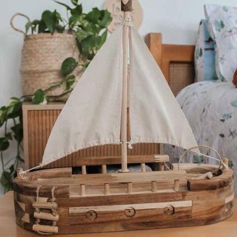 Wooden model boat with a white sail on a wooden surface, surrounded by plants and decor.
