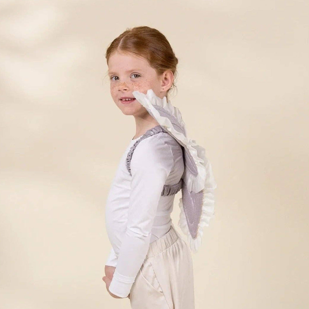 A young girl with light brown hair tied back is shown in profile wearing a white long-sleeve top with a gray ruffled collar, cream wide-leg pants, and white shoes, standing against a warm beige studio backdrop.