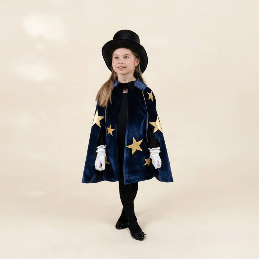 A young girl modeling a magician costume consisting of a navy blue velvet cape with gold stars, a black top hat, white gloves, and black tights, set against a solid beige studio background.