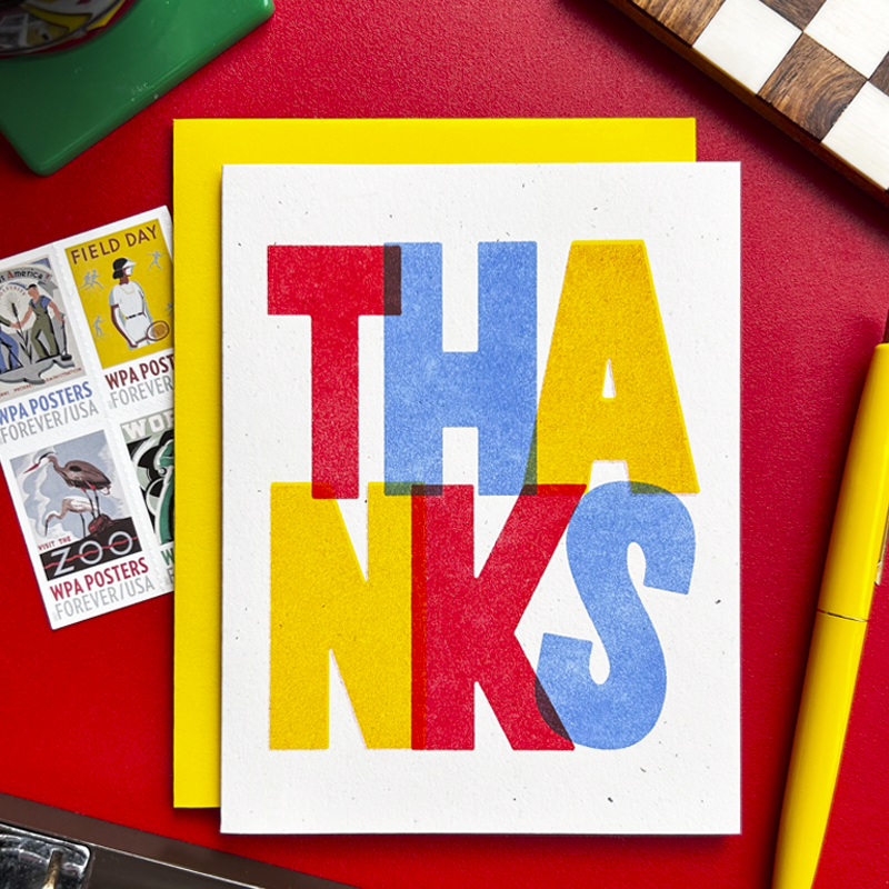 A square white greeting card with large colorful letters spelling THANKS, layered over a bright yellow card, photographed on a red surface with postage stamps and a pen nearby.