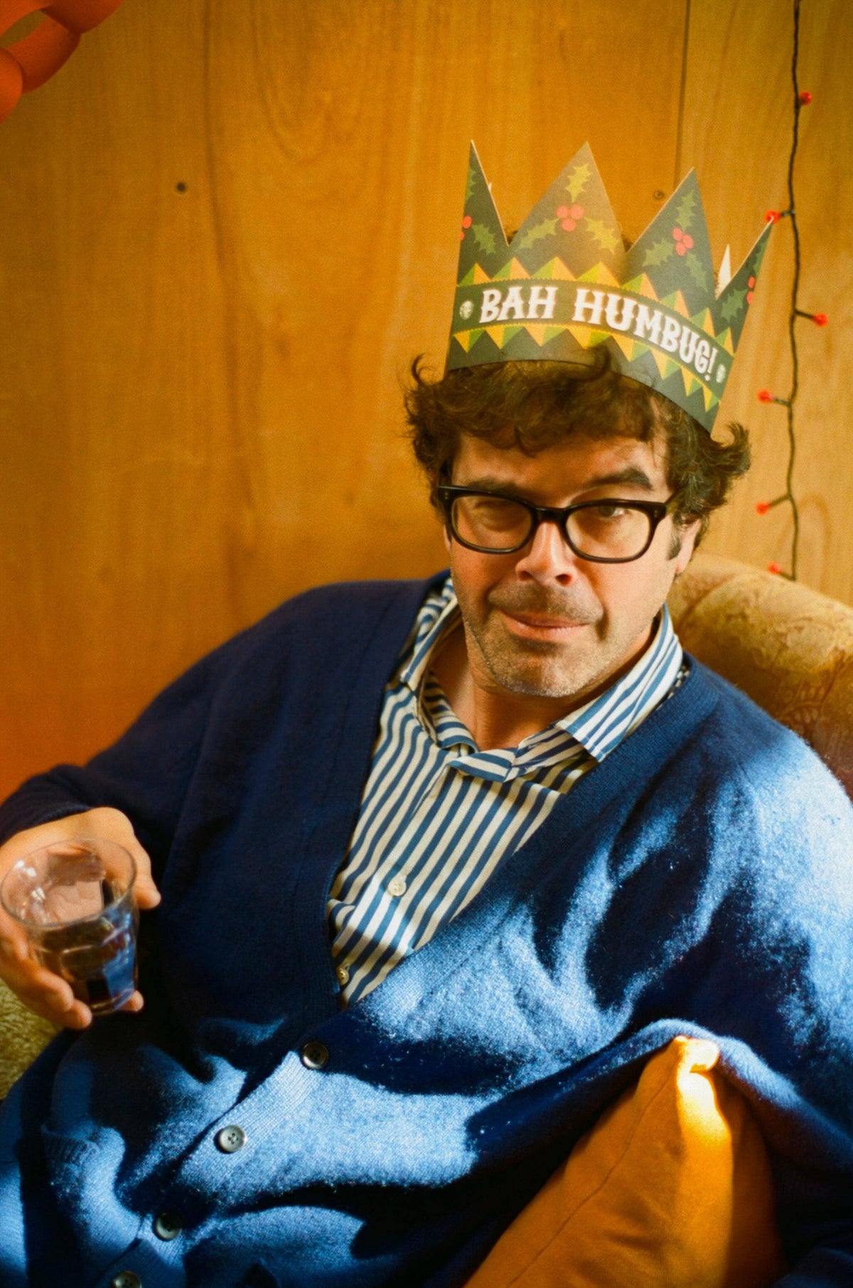 A man wearing thick black glasses, a blue cardigan, and a striped shirt sits in a warm indoor setting, wrapped in a blue blanket. He is wearing a paper crown that reads 'BAH HUMBO!'.