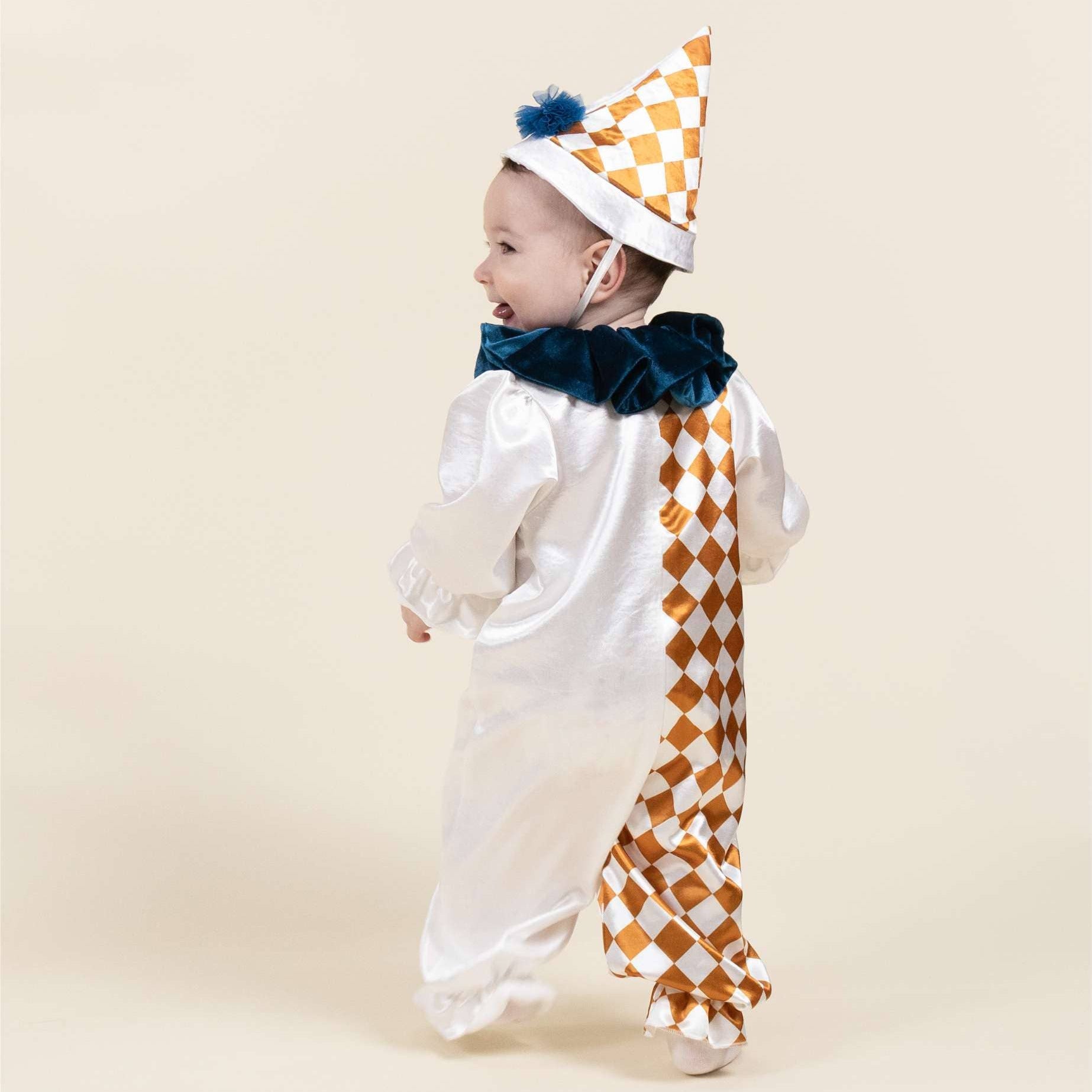 A baby wearing a white clown costume with a gold and white checkered pattern on the back and hat, and a dark blue ruffled collar, standing against a solid beige background.