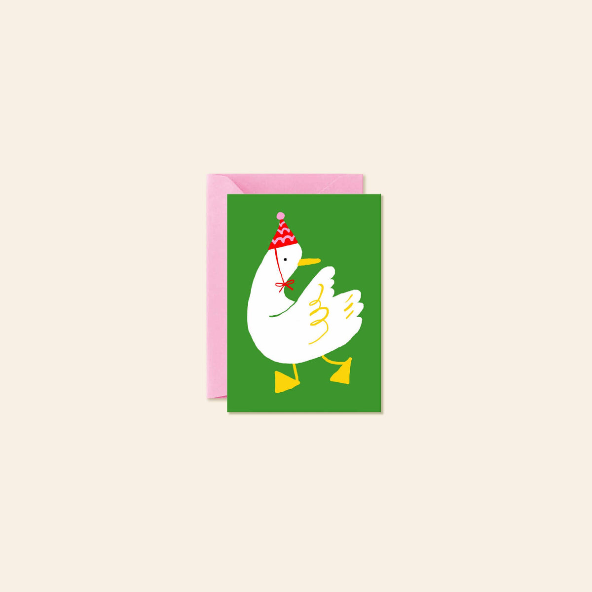 A greeting card with a pink envelope behind it, featuring a white cartoon duck wearing a red and white party hat on a green background, set against a light beige backdrop.