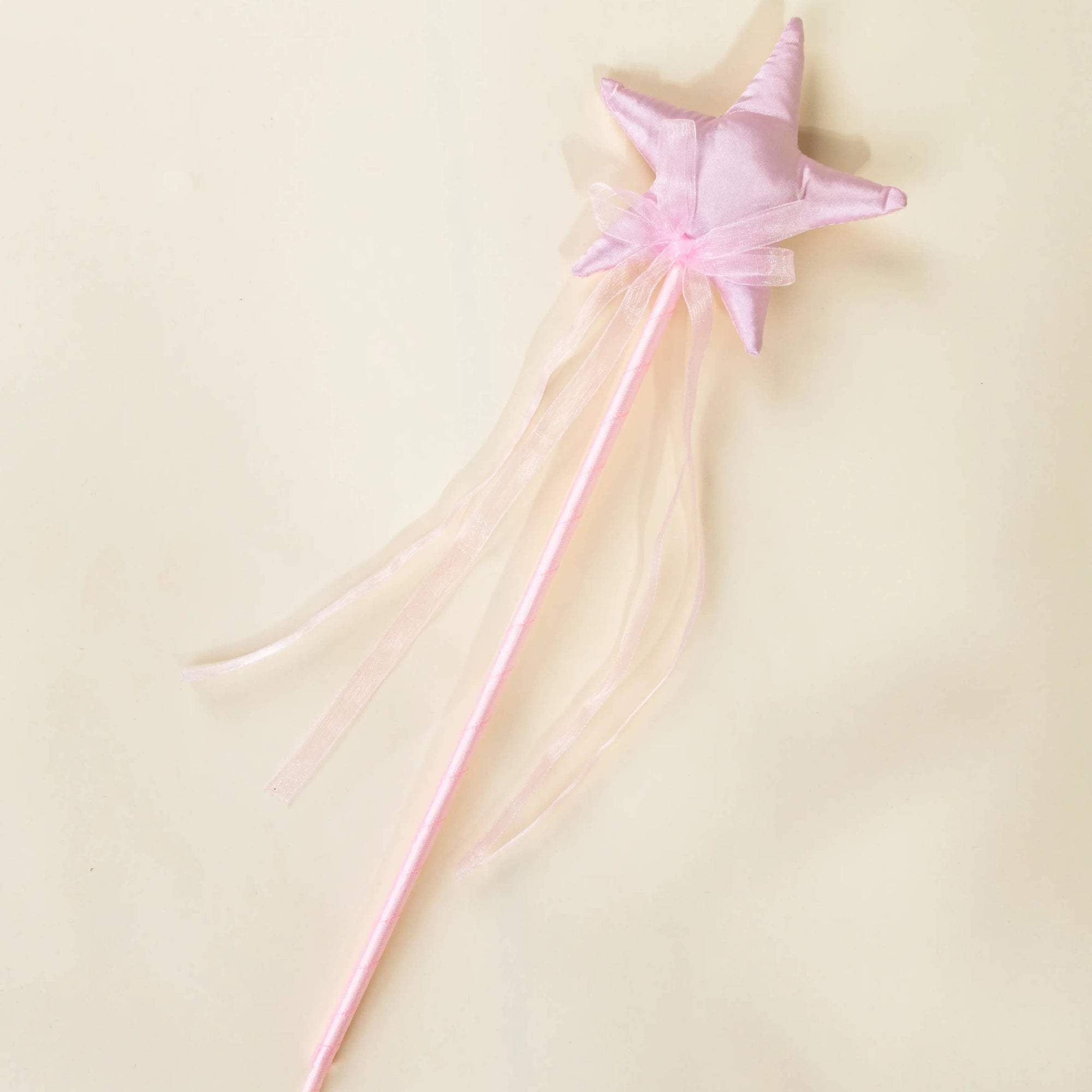 A pink star-shaped magic wand with a long pink handle and sheer pink ribbon tails, photographed on a plain light beige background.