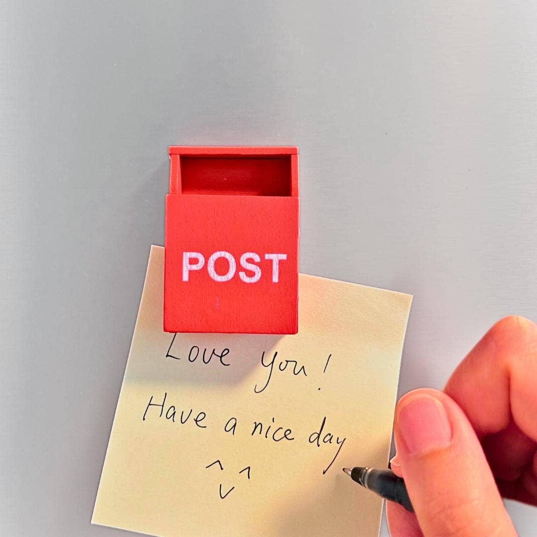 A small red post box with the word POST in white letters, attached to a beige sticky note that reads 'Love you! Have a nice day' with a hand holding a pen near it, against a light gray background.