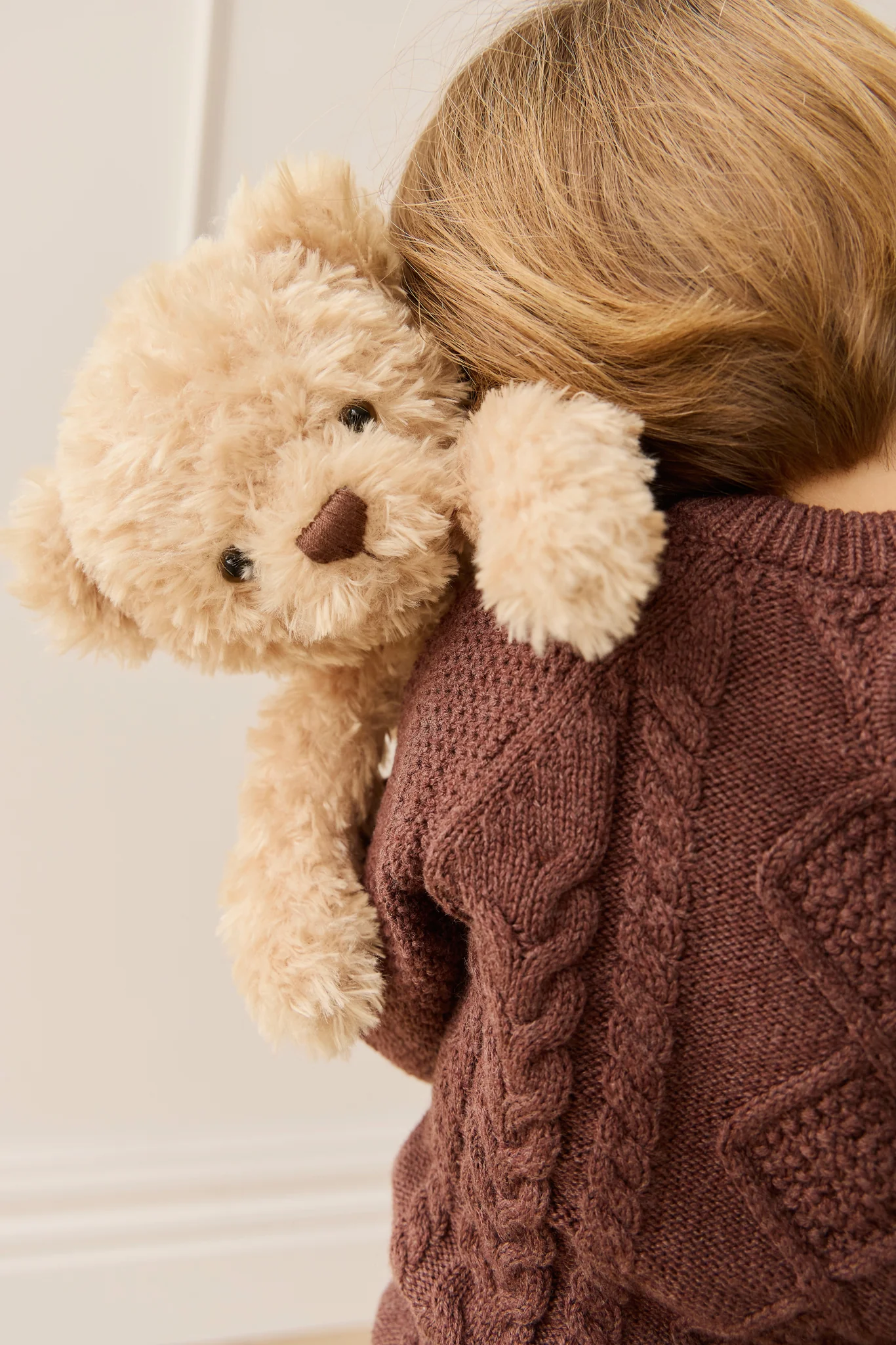 Person wearing a brown sweater holding a beige teddy bear against a neutral background