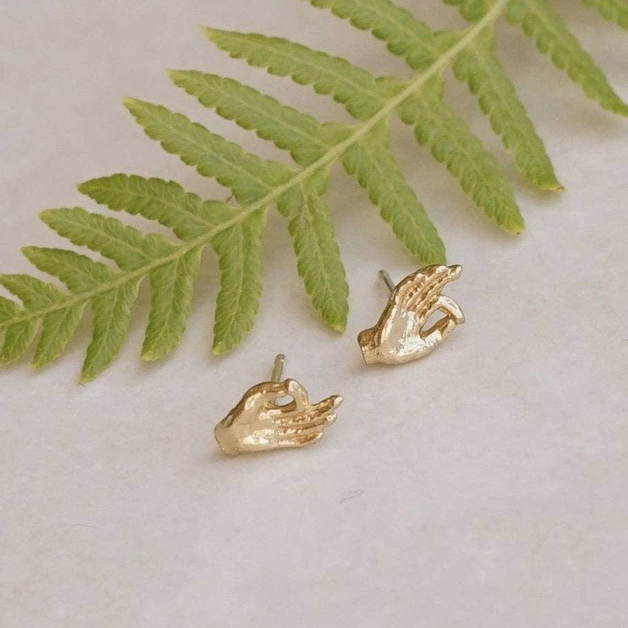Gold hand-shaped earrings on a light surface with a green leaf