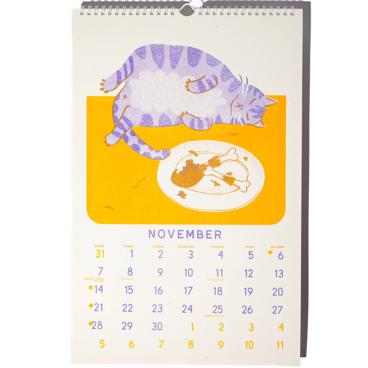 A spiral-bound November desk calendar featuring a stylized illustration of a purple-striped cat lying over a yellow-orange block above a plate of food, with the word 'NOVEMBER' and numbered days below on a white page.