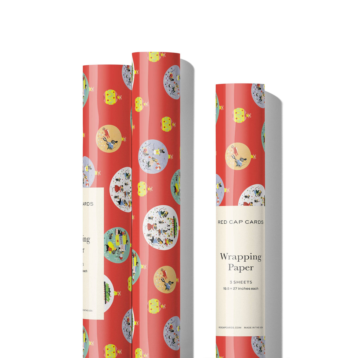 Three rolls of wrapping paper on a white background, featuring a red base with a whimsical pattern of yellow ducks, green birds, and round illustrated medallions. One roll is standing upright on the right with a cream paper band labeled 'Red Cap Cards Wrapping Paper'.