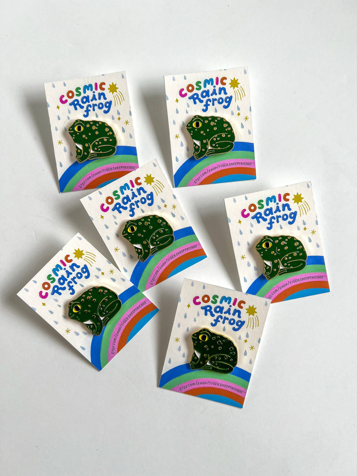 Five 'Cosmic Rain Frog' pins on their packaging with a white background