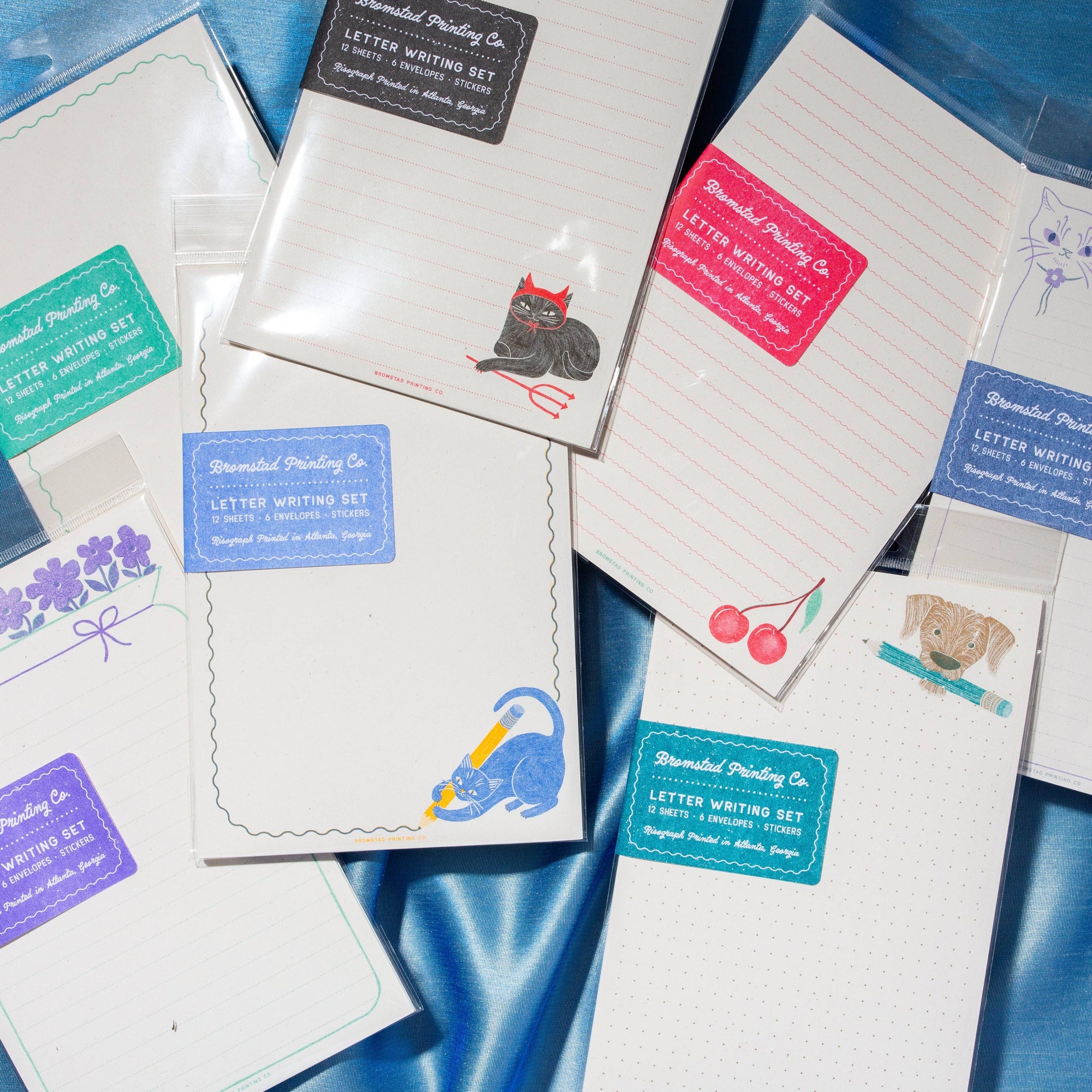 A flat-lay arrangement of multiple letter writing sets from 'Handmade Printing Co.' on a shiny blue satin background.
