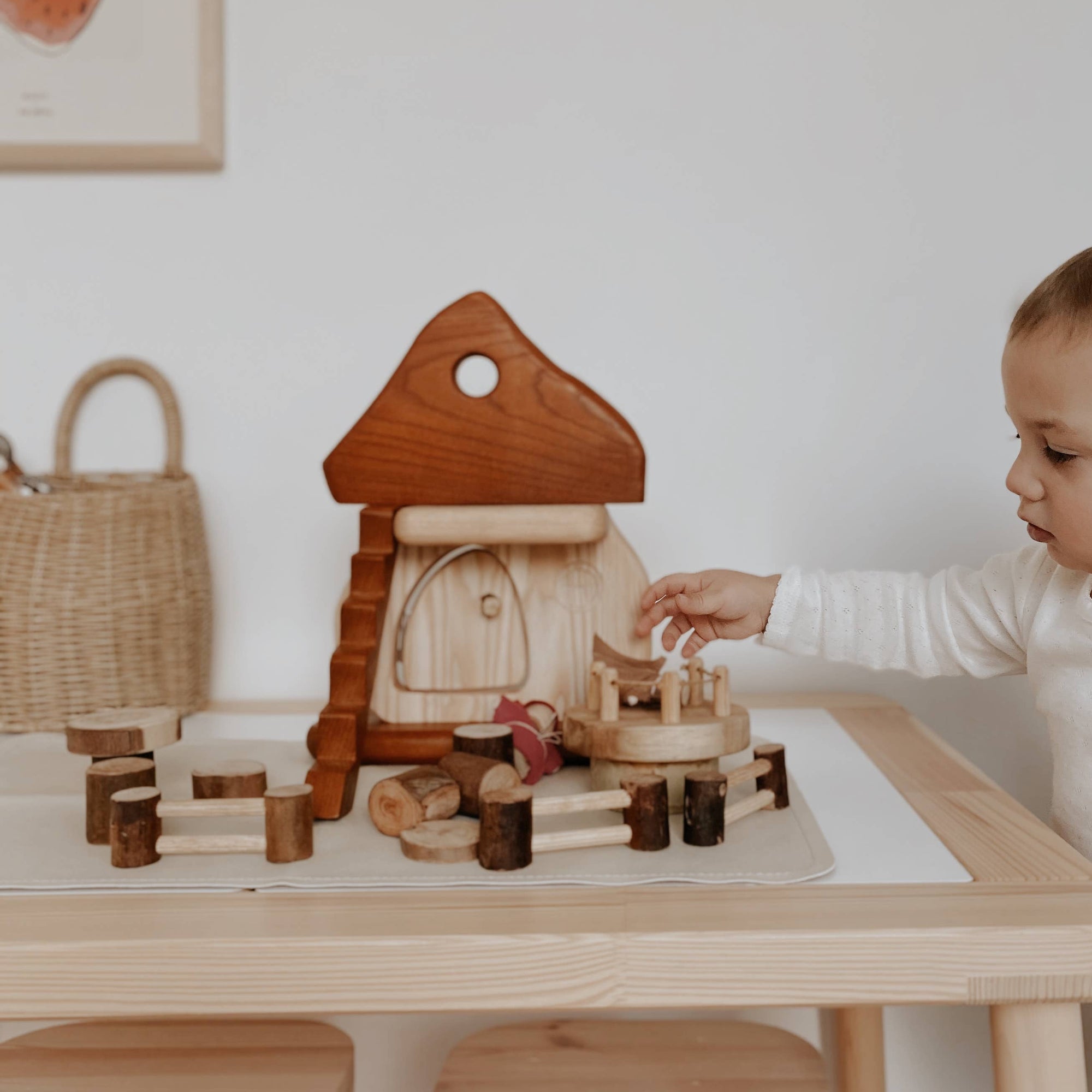 Child playing with a wooden toy set at a table in a room with a white wall and framed picture.