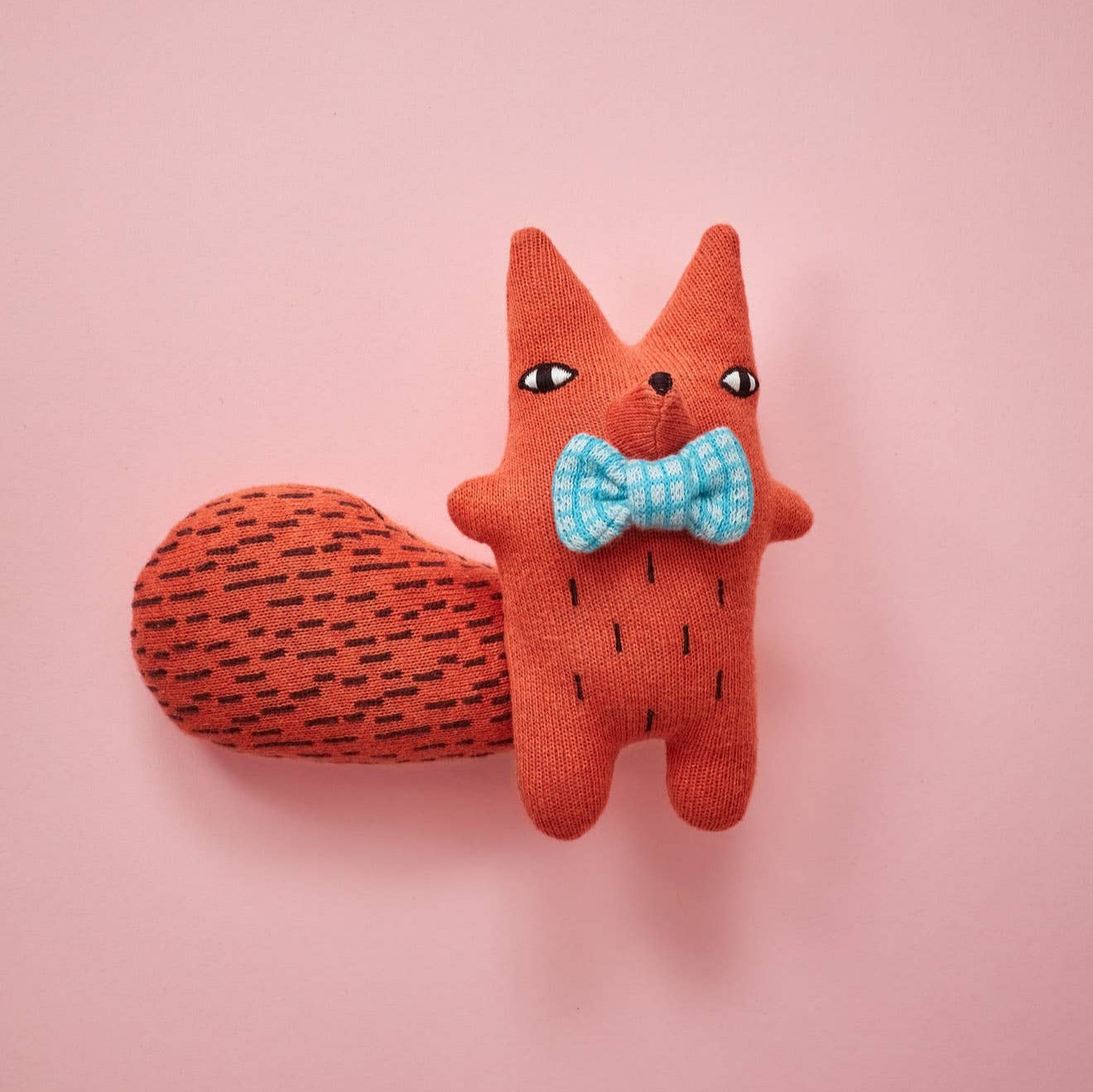 A plush toy representing a squirrel with a bow tie, primarily in orange color, presented against a pink background.