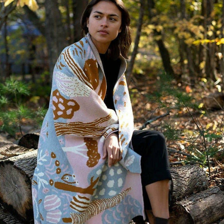 Woman sitting on a log in a forest, wrapped in a colorful blanket with nature-themed patterns.