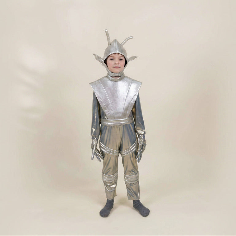 A child wearing a shiny silver alien or sci-fi costume with a helmet, shoulder armor, gloves, and matching pants, standing against a plain beige studio background.