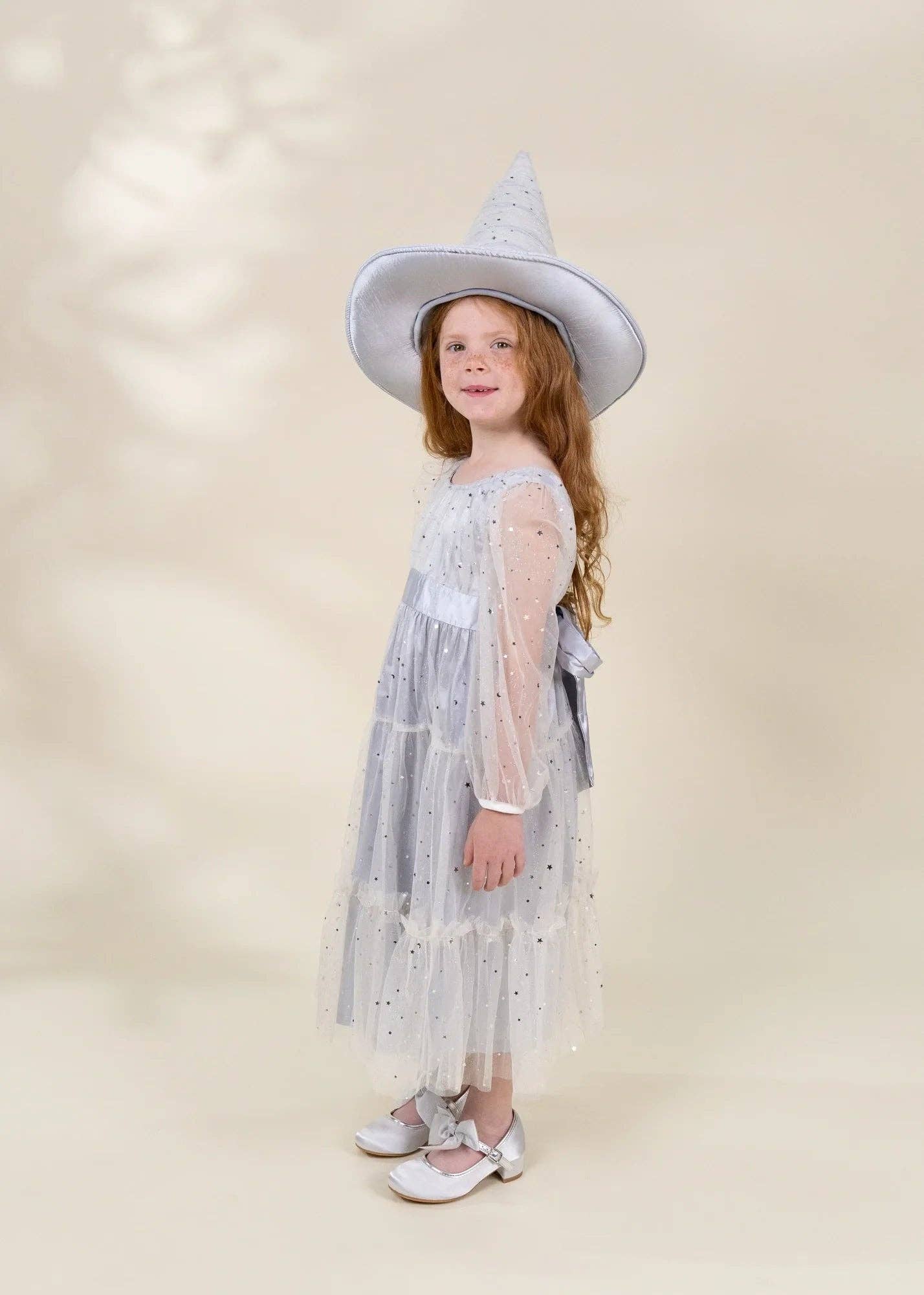 A young girl with long light brown hair stands in profile wearing a light blue-gray witch-style costume dress with sheer sleeves, a pointed hat, and silver shoes against a warm beige studio background.