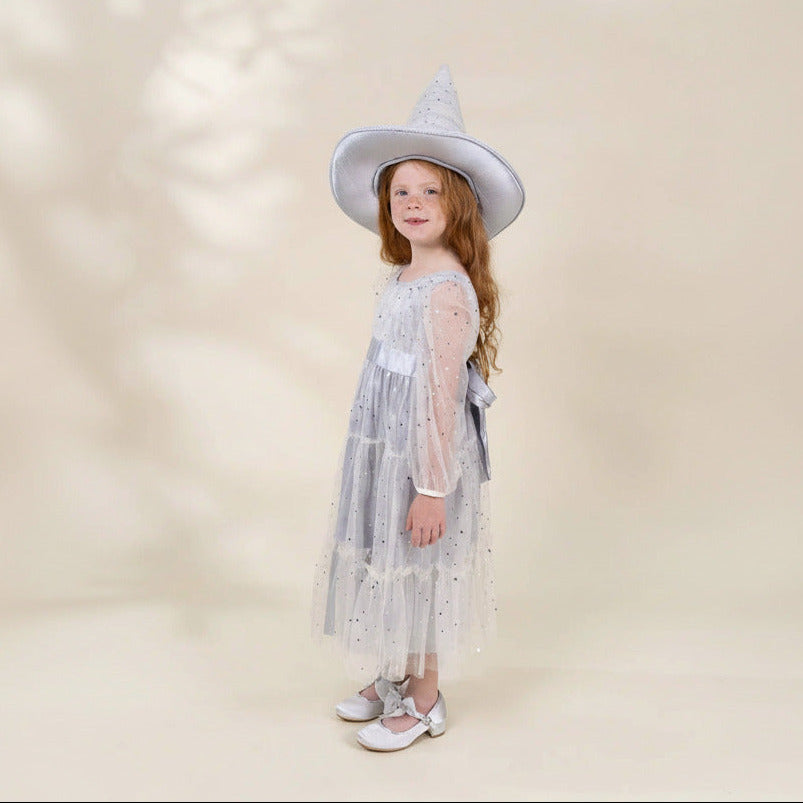 A young girl with long light brown hair stands in profile wearing a light blue-gray witch-style costume dress with sheer sleeves, a pointed hat, and silver shoes against a warm beige studio background.