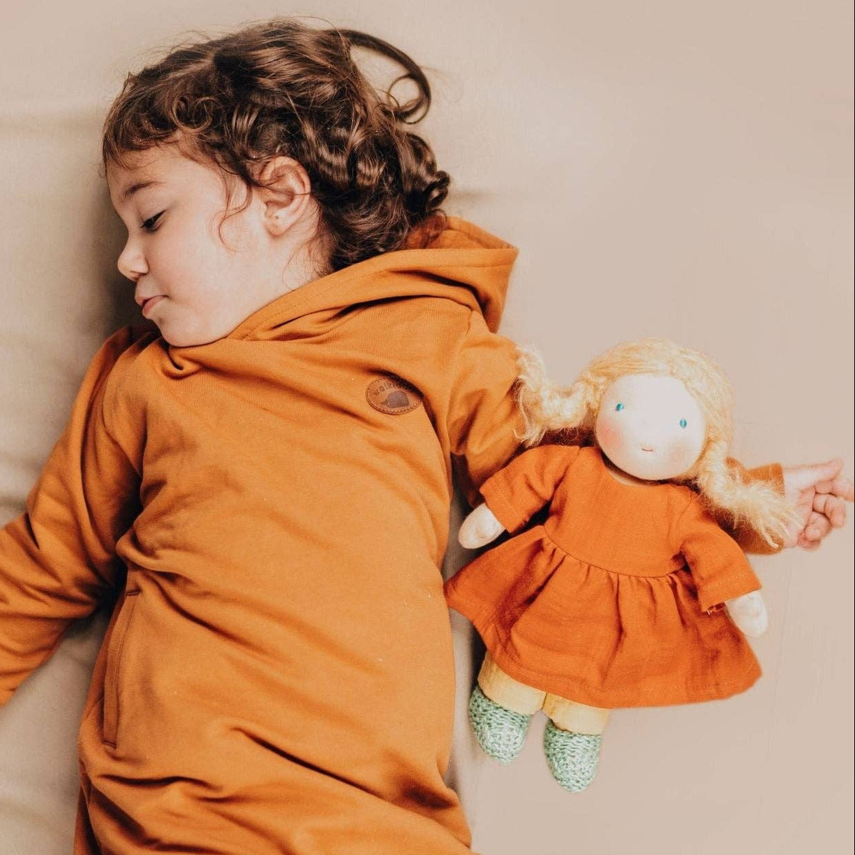 A child wearing a burnt orange hooded sweatshirt and matching pants lies on a beige bed while holding a small blonde doll dressed in an orange dress.