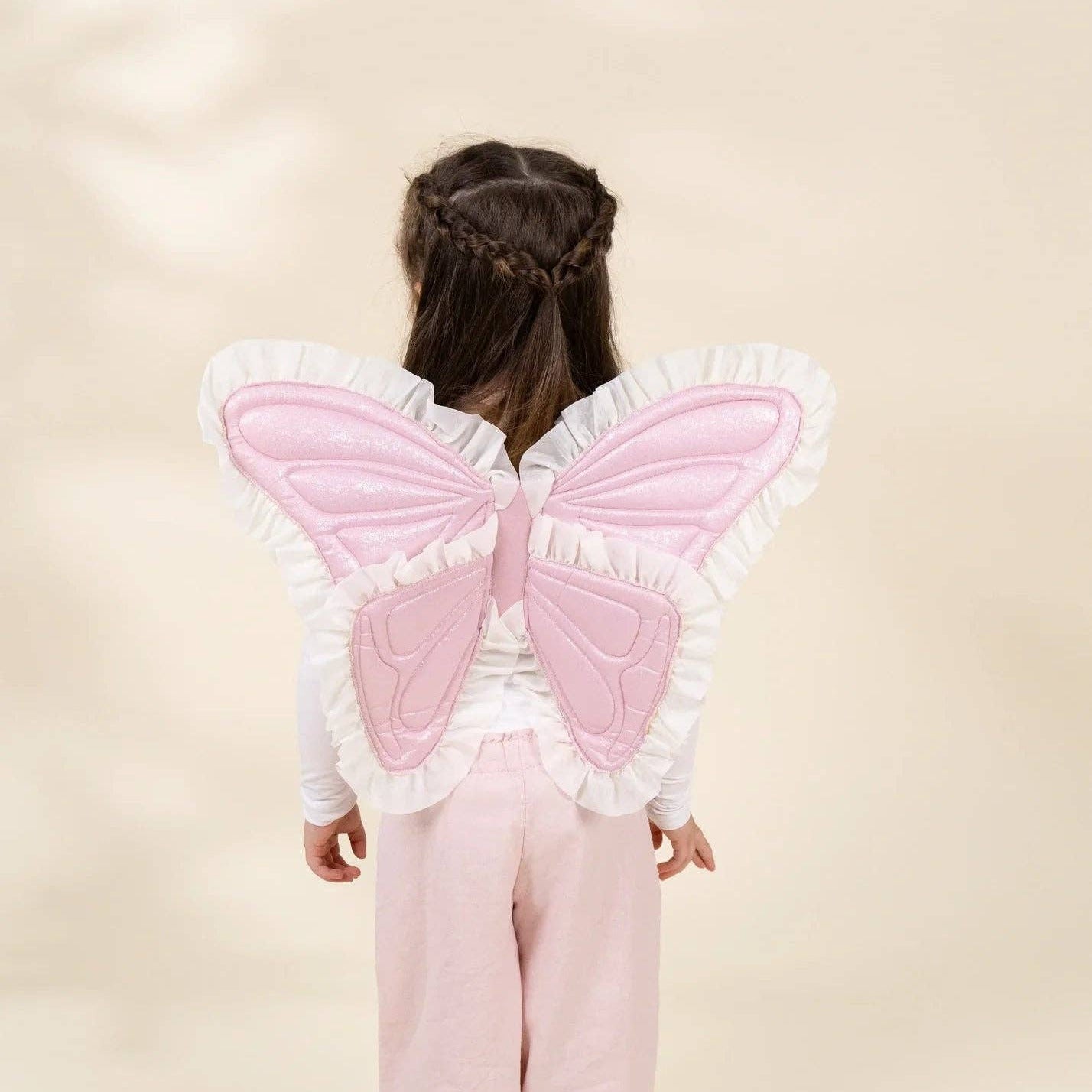 A child viewed from behind wearing a pink and white fairy costume with large quilted butterfly wings and white ruffled trim, paired with light pink pants, against a soft beige studio background.