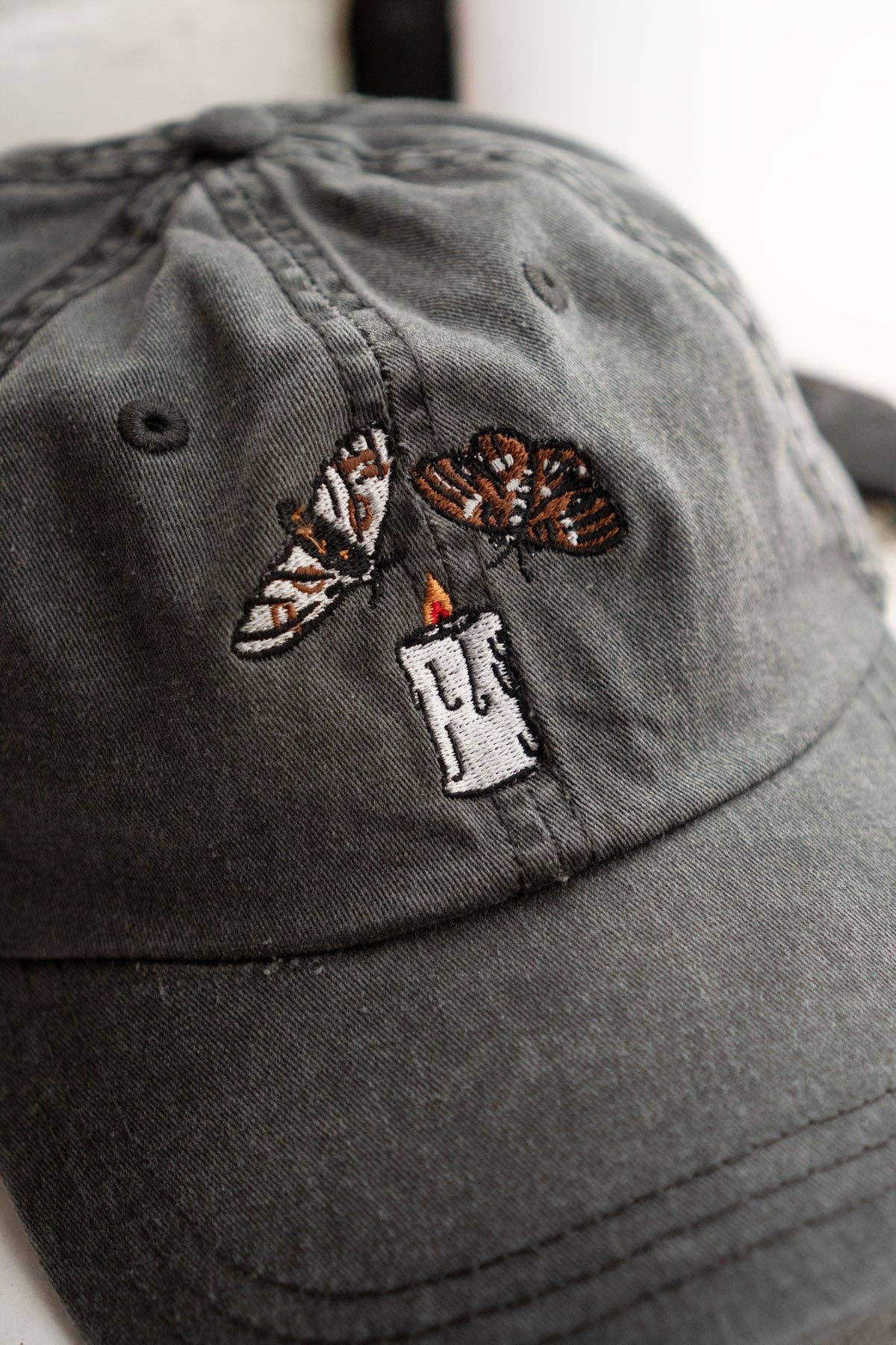 Gray cap with embroidered designs of a candle and moths on a neutral grey background.