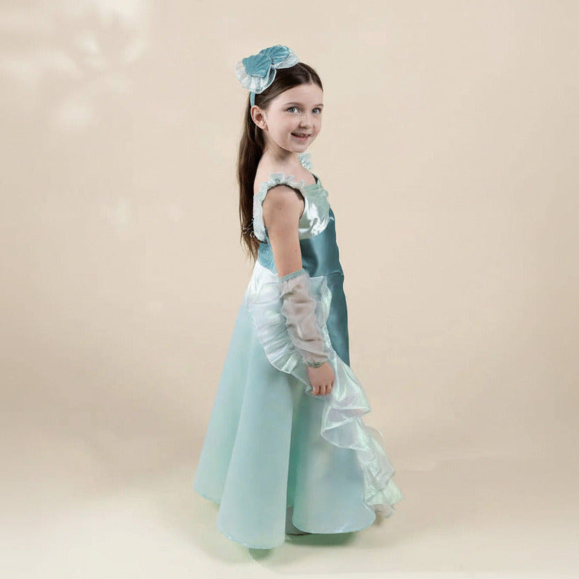 A young girl modeling a light blue and white princess-style dress costume with matching blue flower headband, standing against a plain beige studio background.