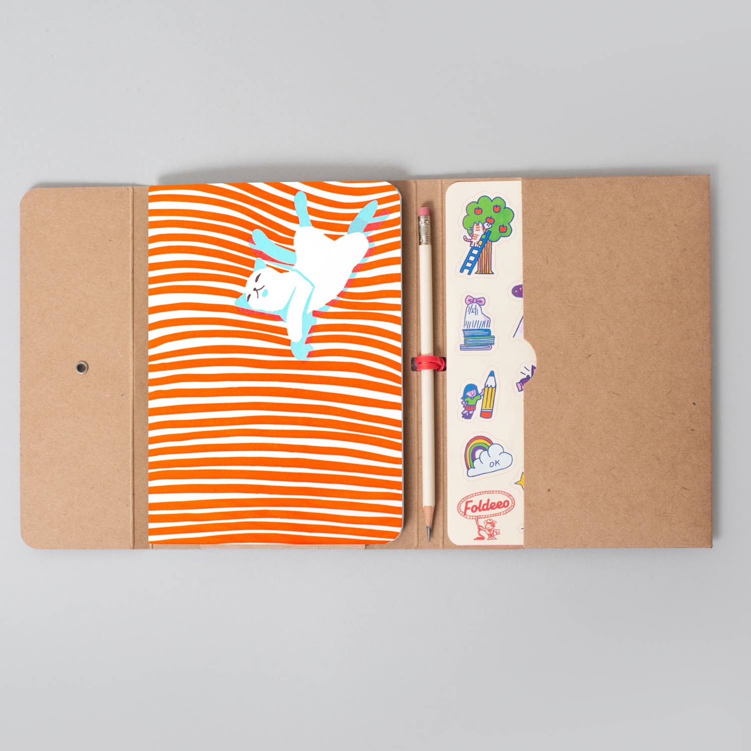 A kraft paper notebook cover opened flat on a light gray background, showing an orange-and-white striped cover with a white unicorn illustration, a pencil in a side pocket, and a small red band labeled Foldables.