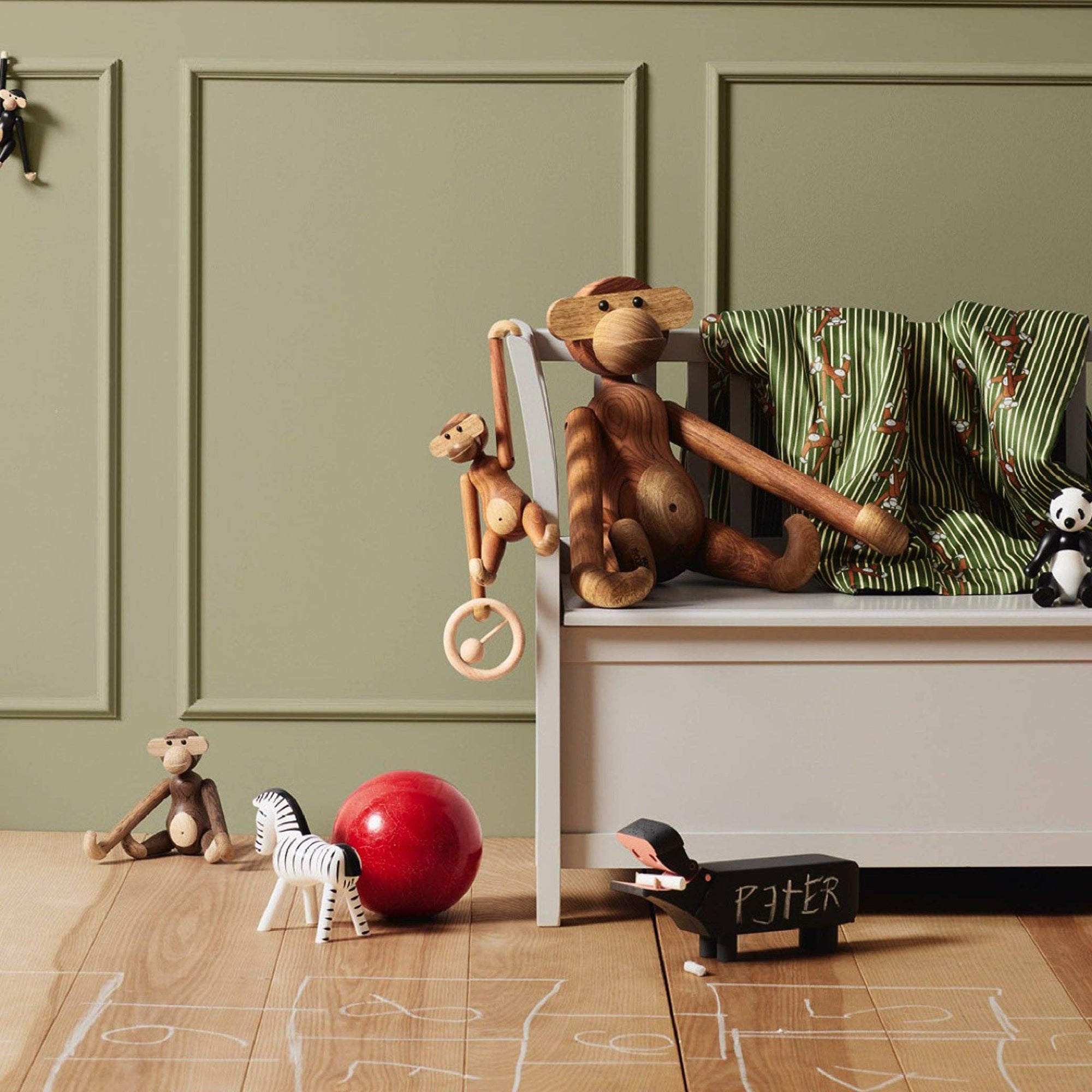 Toy monkey on a seesaw with other toys on a wooden floor against a green paneled wall.