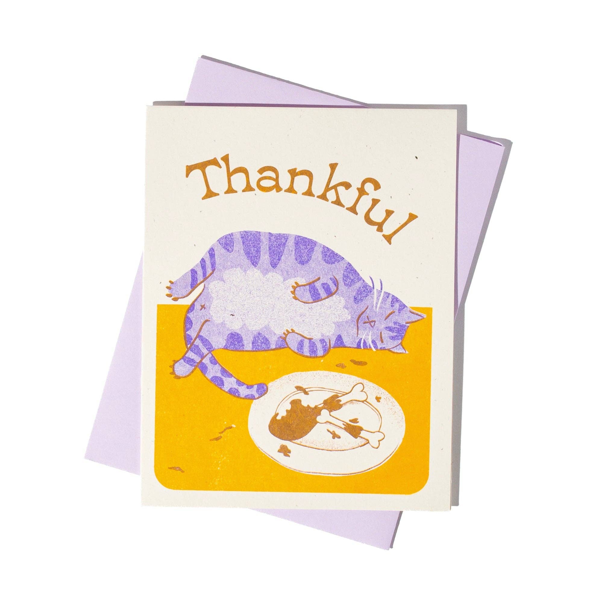 A greeting card with a lavender envelope behind it, featuring the word 'Thankful' in brown above an illustration of a purple-striped cat lying on a yellow surface next to a plate of food.