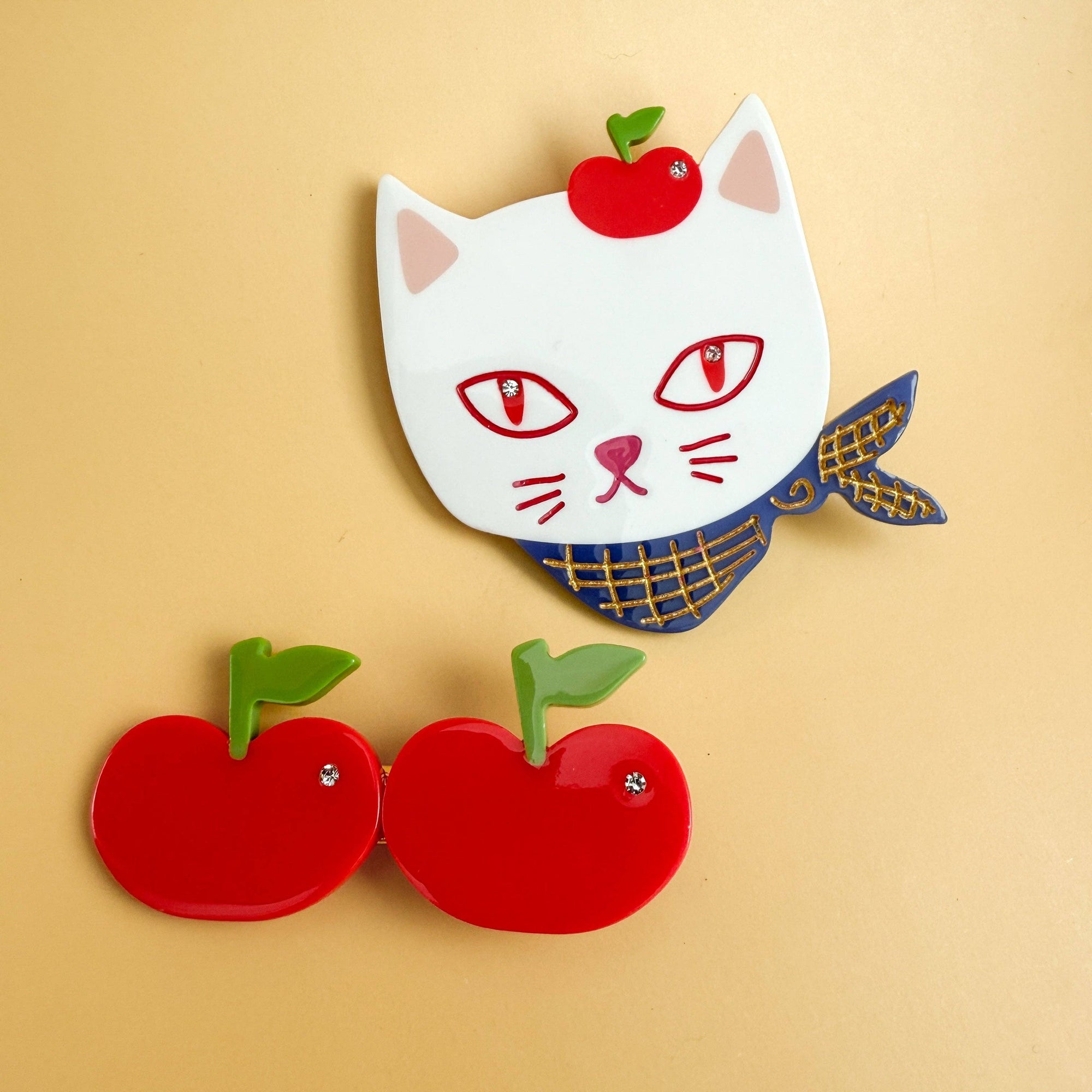 Cat-shaped brooch with scarf and red apples on a beige background