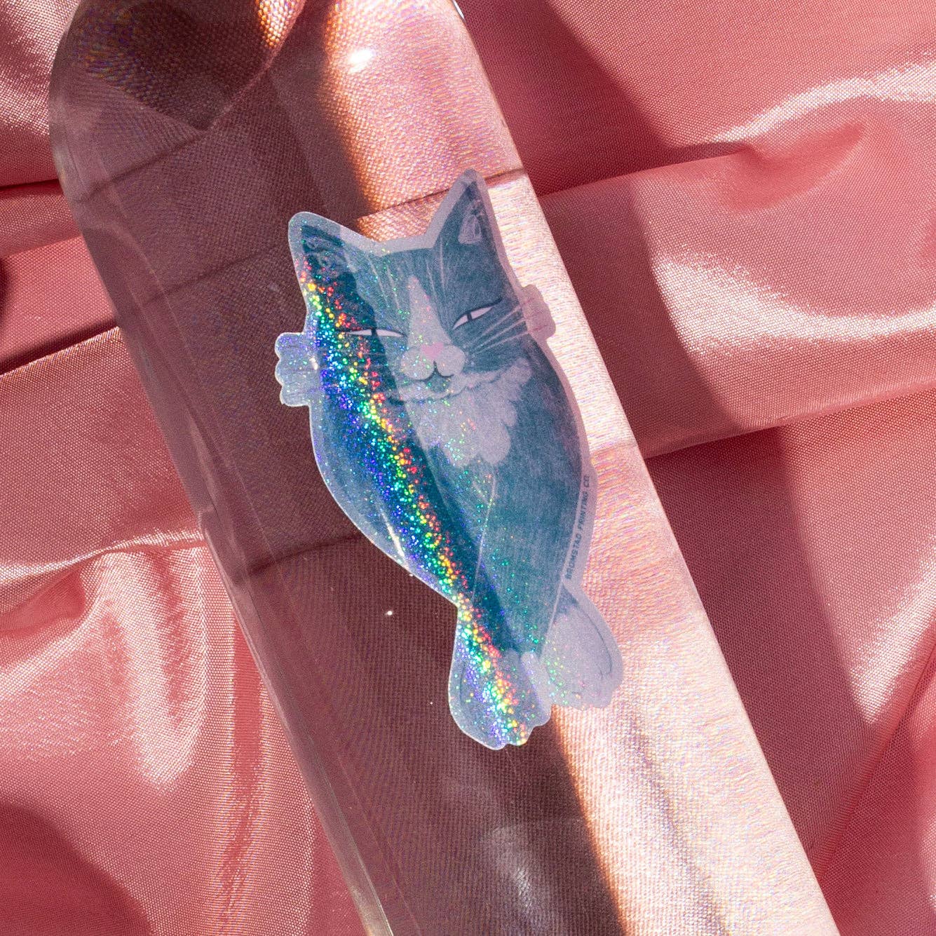 A die-cut sticker of a blue cat with white details and a holographic iridescent stripe, shown resting on a pink satin fabric background.