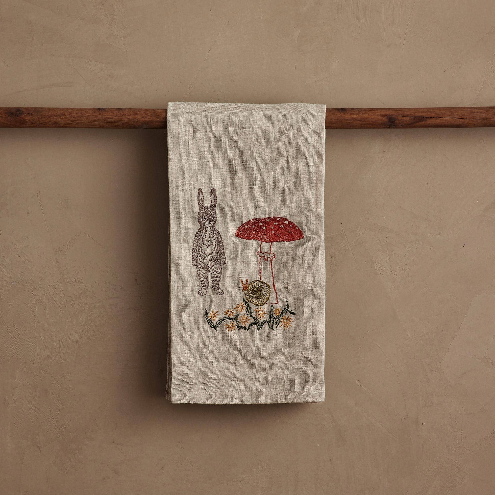 Textile with embroidered design of a rabbit and mushroom hanging on a wooden rod against a beige wall.