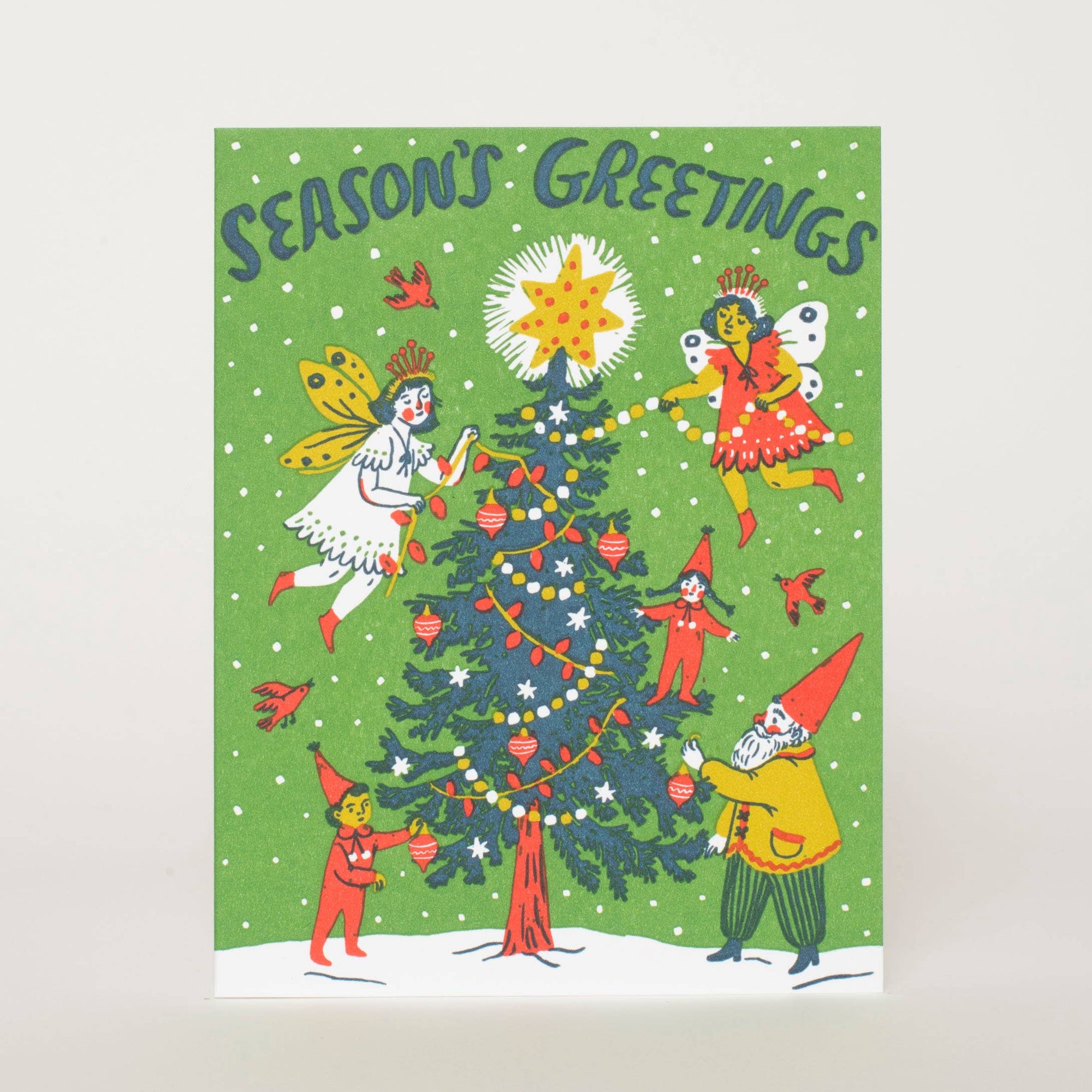 A vintage-style Christmas greeting card featuring a green background with the words 'SEASON'S GREETINGS' at the top. The card illustration shows a decorated Christmas tree with a star topper, small holiday figures, birds, and snowflakes in red, yellow, white, and blue tones.