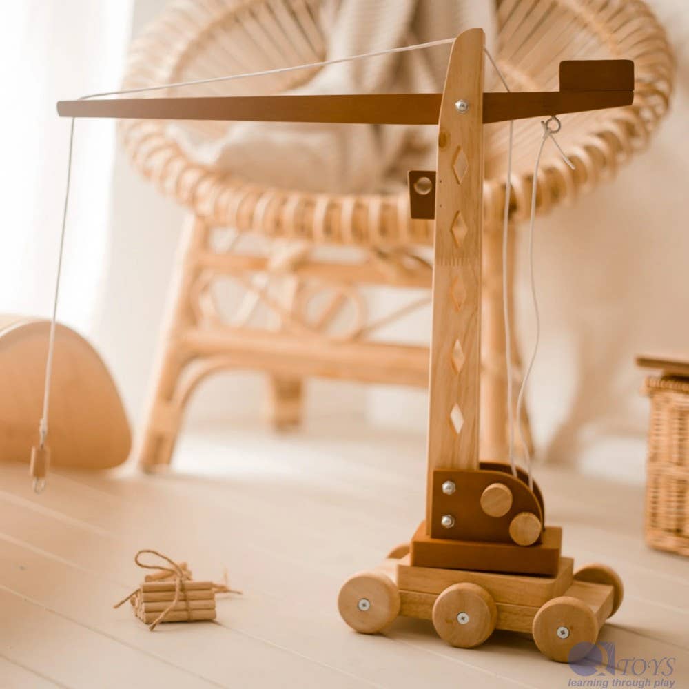 A wooden toy crane or catapult-style children's toy with wheels, shown on a light floor in a softly blurred indoor setting with a wicker chair in the background.