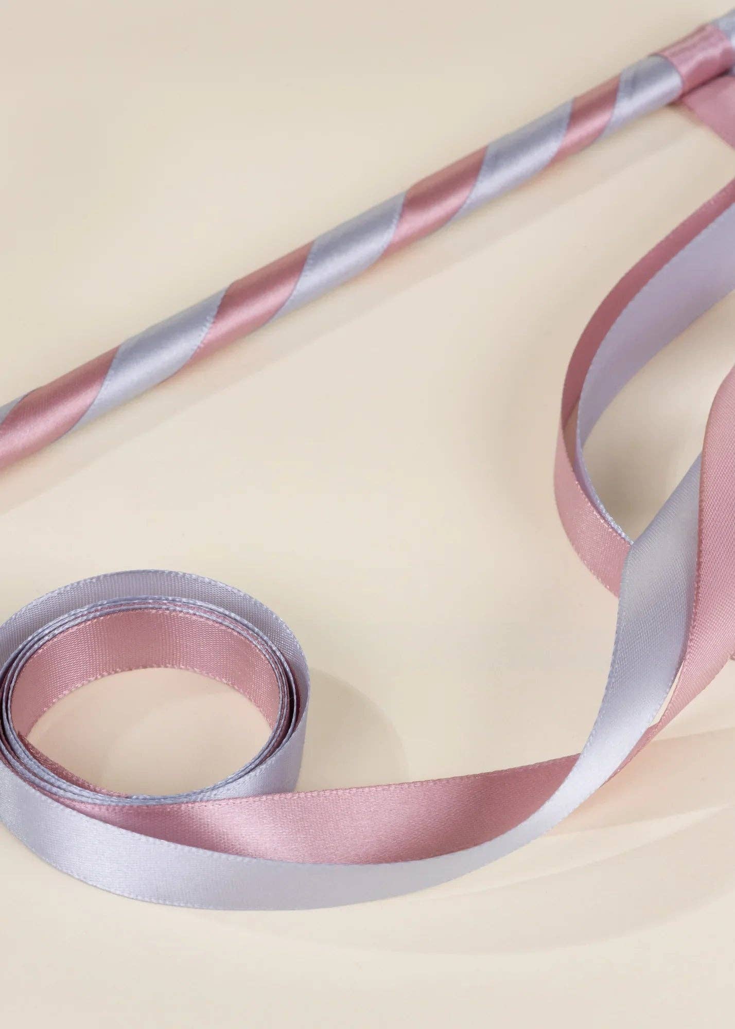 A close-up of a satin ribbon with alternating pastel pink and light lavender-blue stripes, shown coiled and draped on a warm cream background.