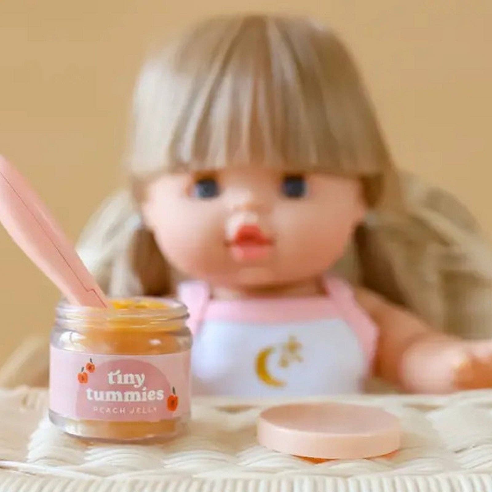 Small jar labeled 'tiny tummies' next to a doll with a pink outfit on a beige surface.