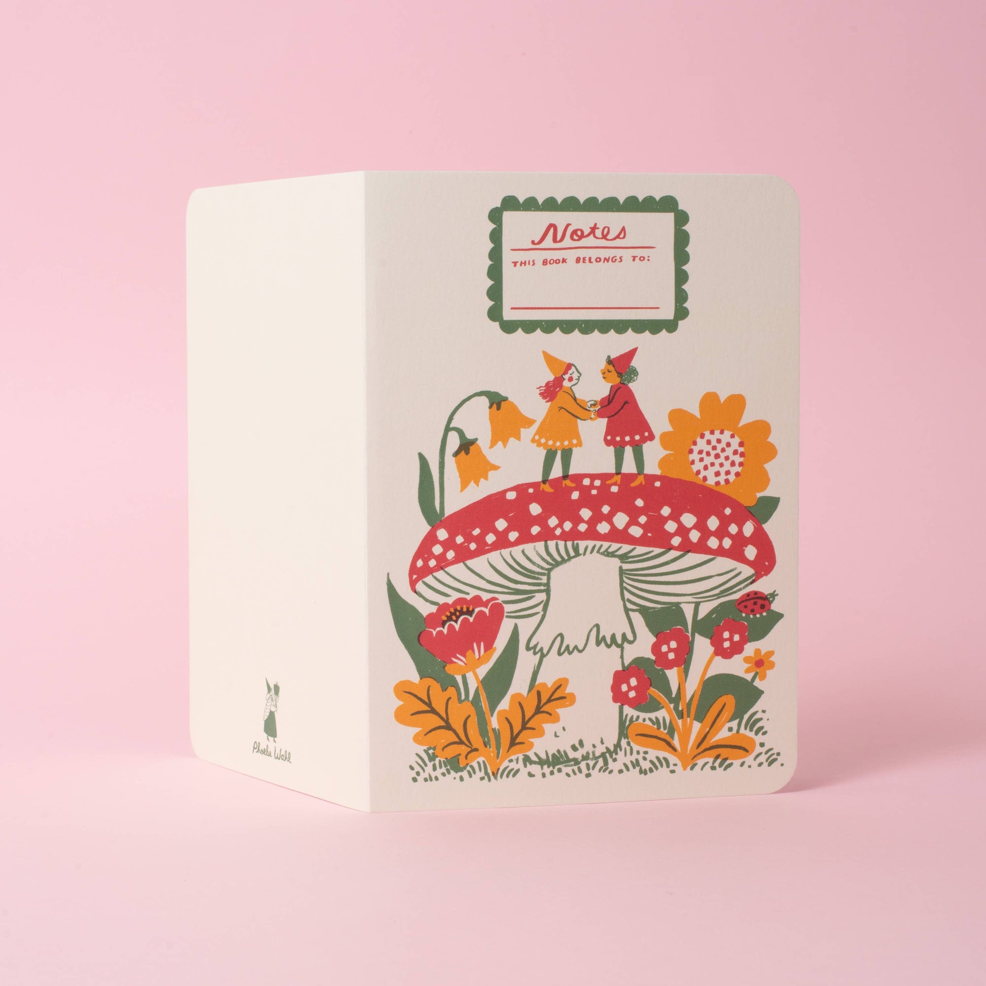 A cream-colored paper notebook or journal with a whimsical folk-art illustration of two fairy-like figures under a large red mushroom, set against a solid pastel pink background. The cover features a green-bordered label at the top reading 'Notes for your favourite pet'.