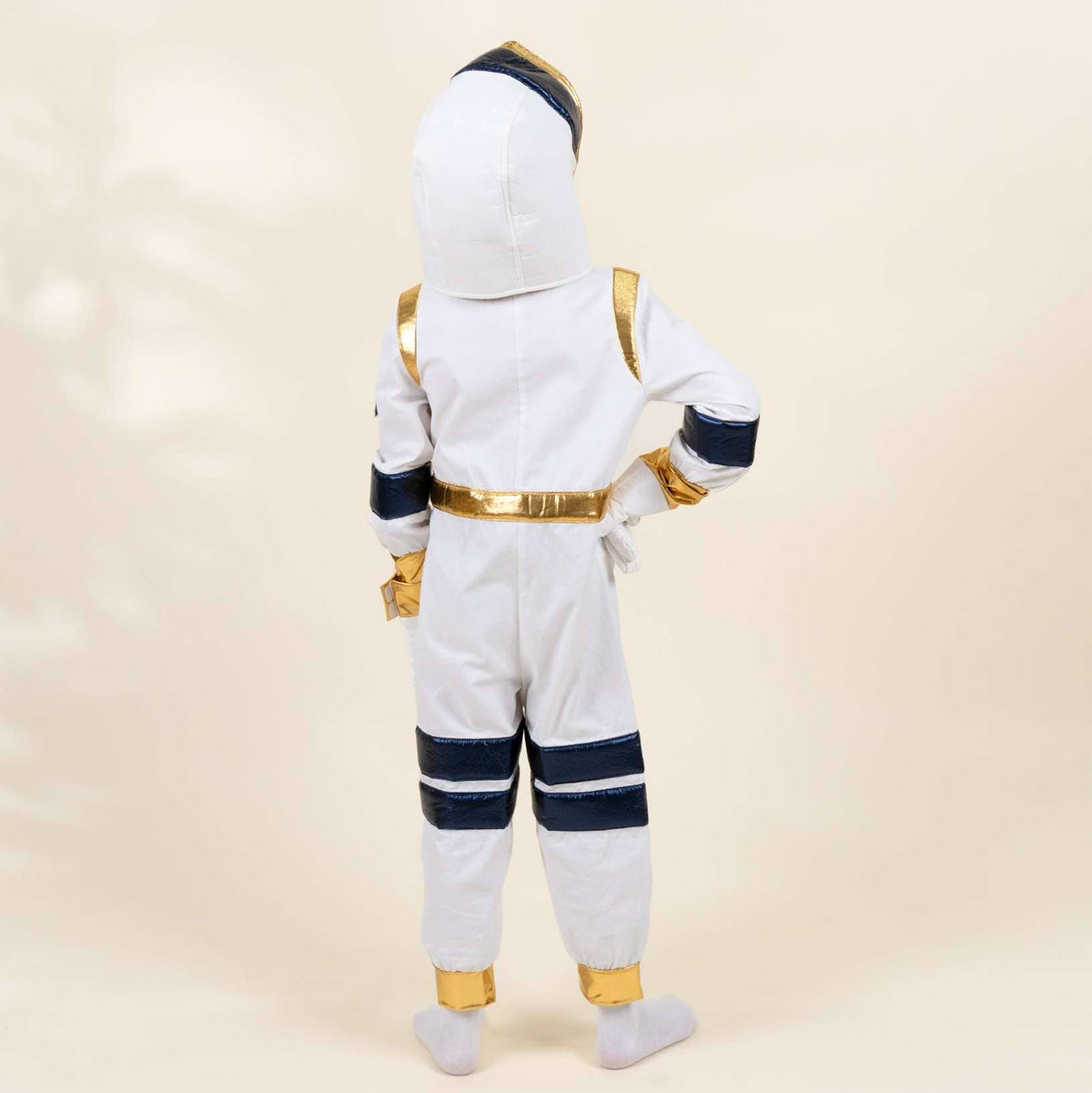 A child wearing a white astronaut costume with gold and navy blue accents, shown from the back against a warm beige studio background.