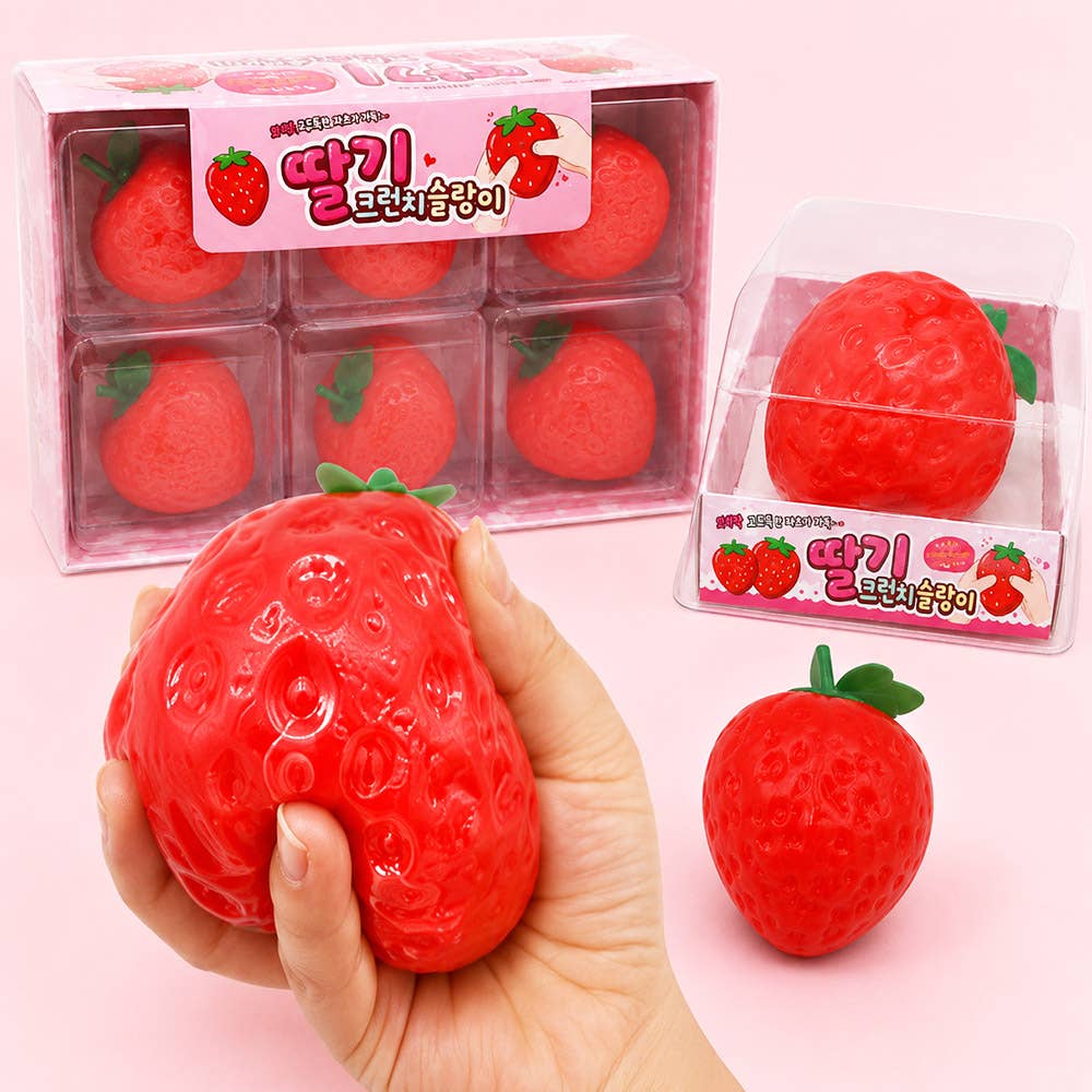 A hand holds a red strawberry-shaped squishy toy in front of its clear plastic packaging, with two more packaged and one loose strawberry squishy toy on a light pink background.