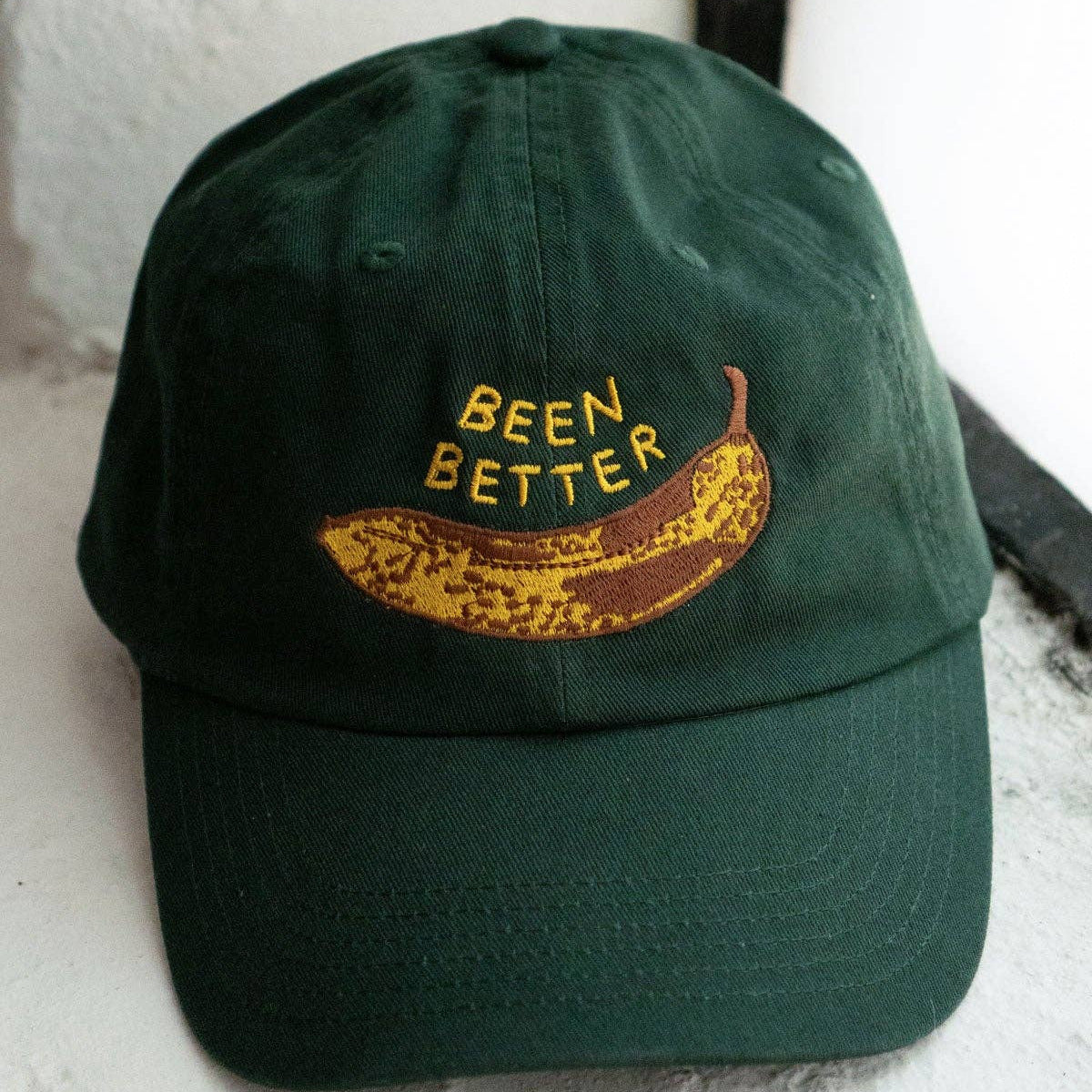 Green cap with a banana graphic and 'BEEN BETTER' text on a white surface.