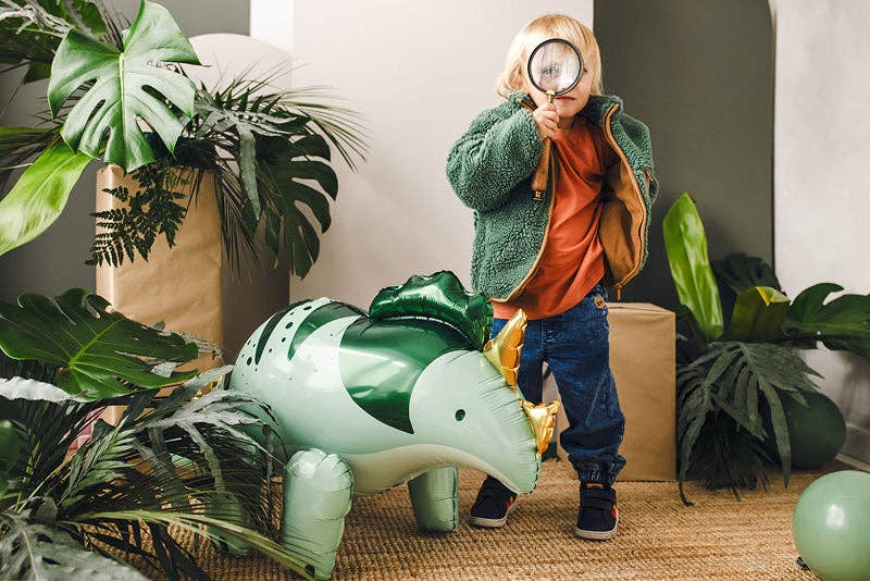 A child wearing a green fleece jacket, orange shirt, and blue jeans stands behind a large inflatable green and white dinosaur balloon, holding a magnifying glass in a decorated indoor setting with plants and cardboard boxes.