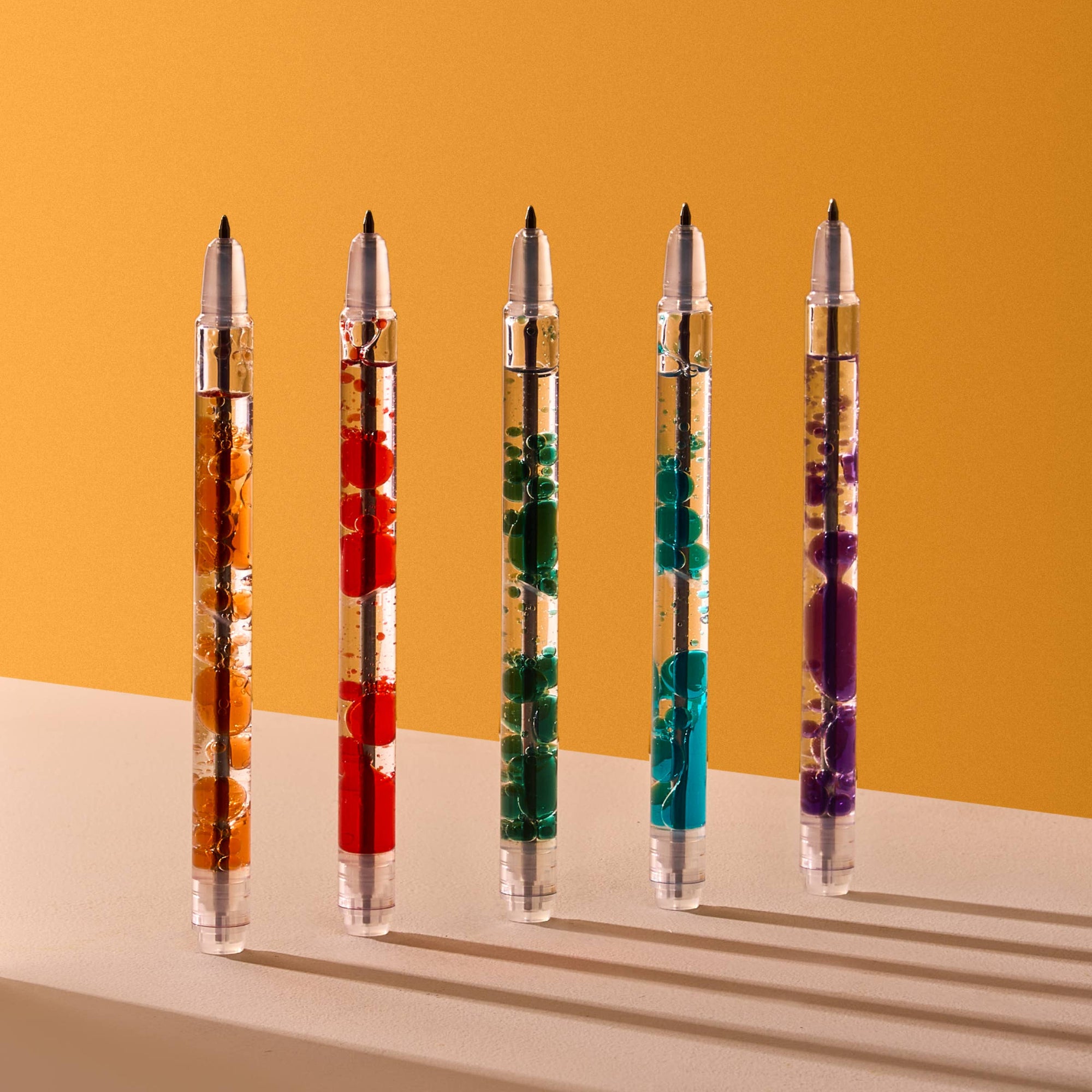 Five decorative pens with colorful interior bubbles on a yellow and beige gradient background