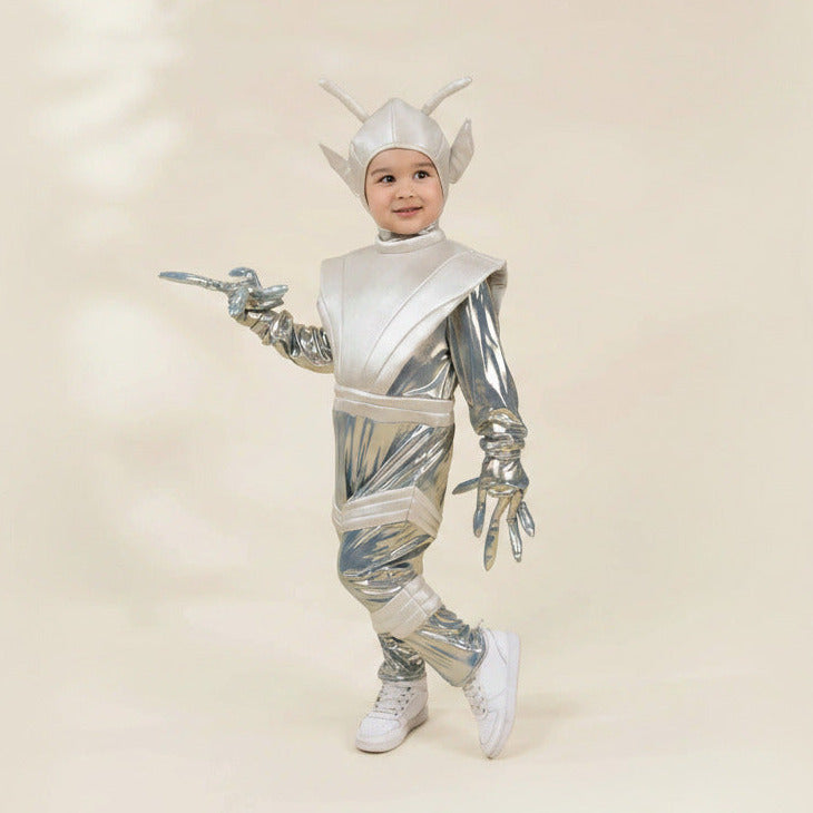 A young child modeling a shiny silver futuristic alien or robot costume with a matching helmet, white gloves, and white sneakers against a plain beige studio background.