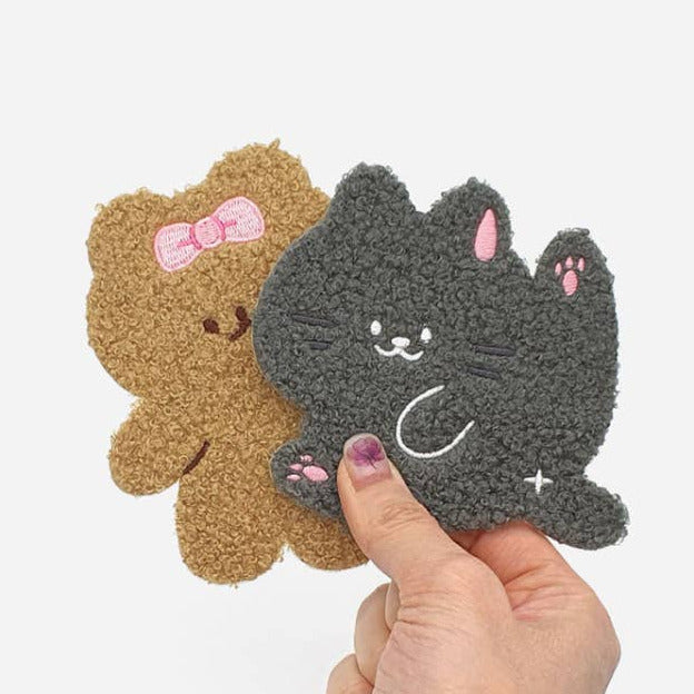 Two plush, textured pet wipes shaped like a brown teddy bear with a pink bow and a dark gray cat with pink ears and paws, held by a hand against a light gray background.