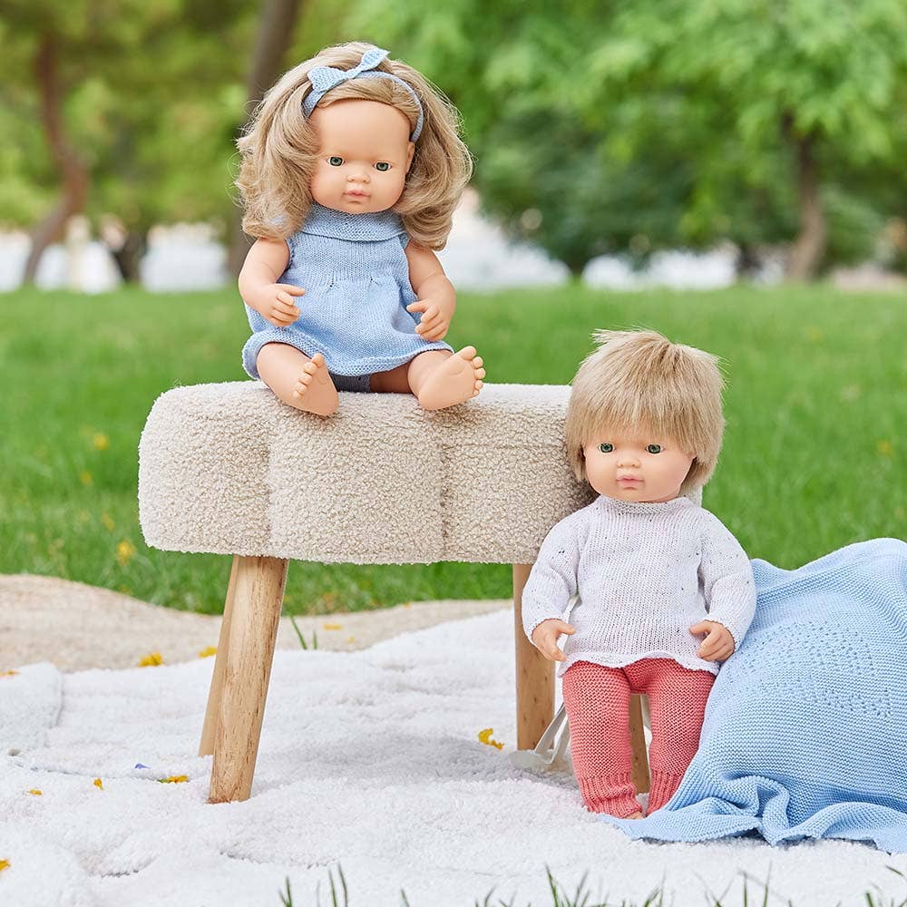 Two dolls sitting on a bench outdoors with greenery in the background