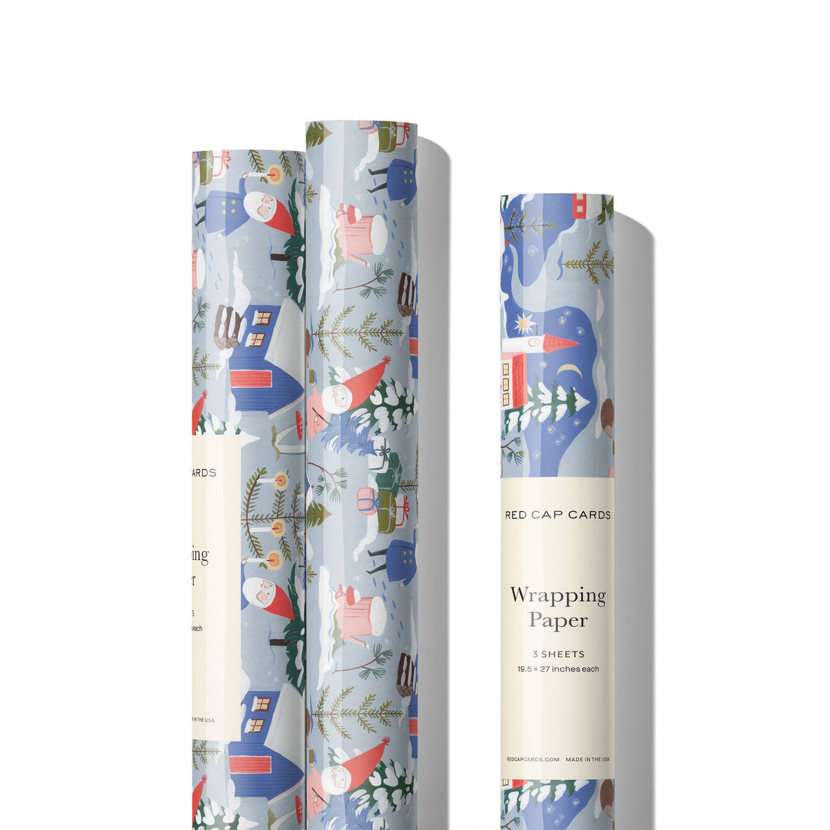 Three rolls of wrapping paper with a whimsical winter village pattern, shown on a clean white background. The pattern features red and pink gnomes, blue houses, and green trees on a light blue-gray background.