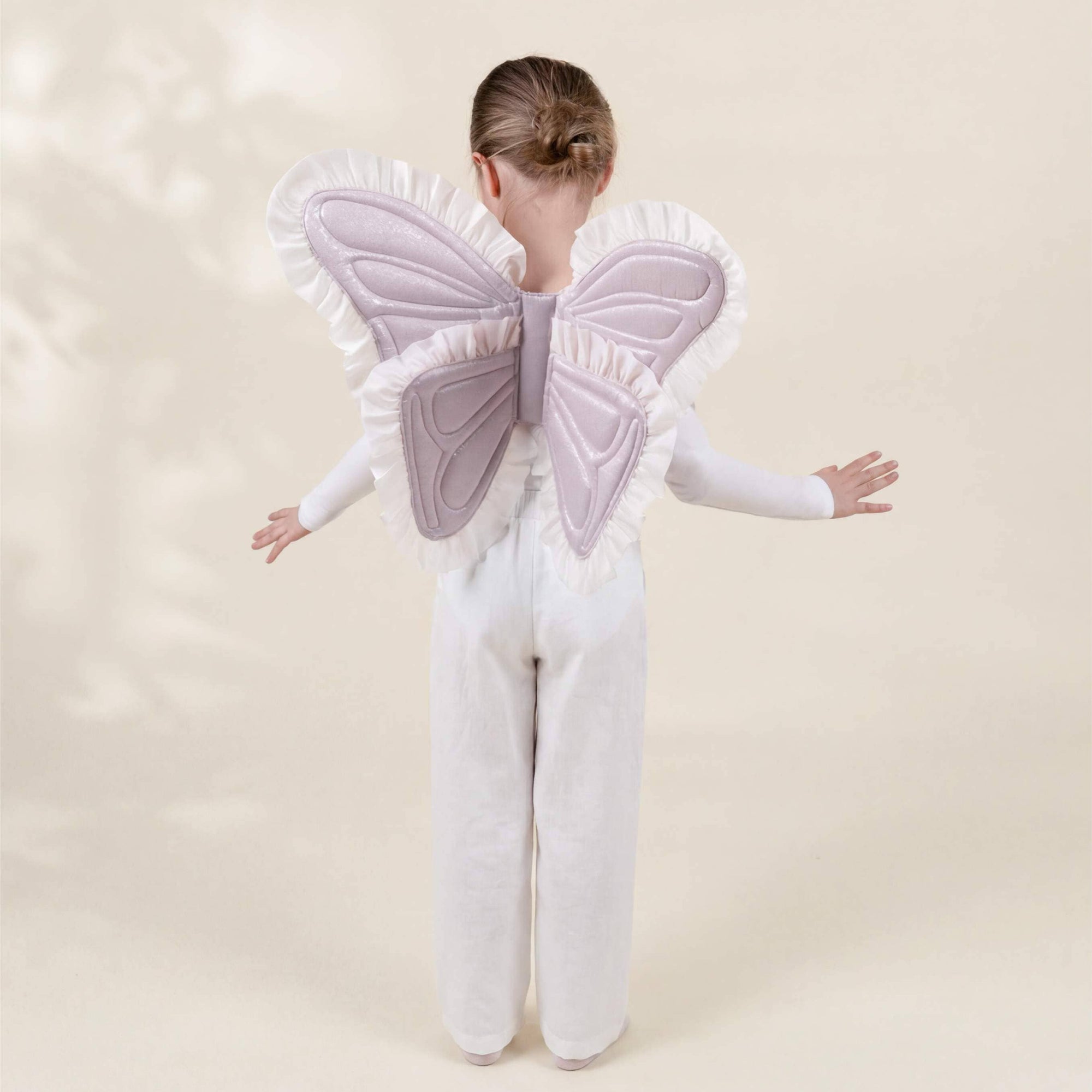 A child viewed from behind wearing a white angel costume with large lavender-pink ruffled wings against a soft beige studio background.